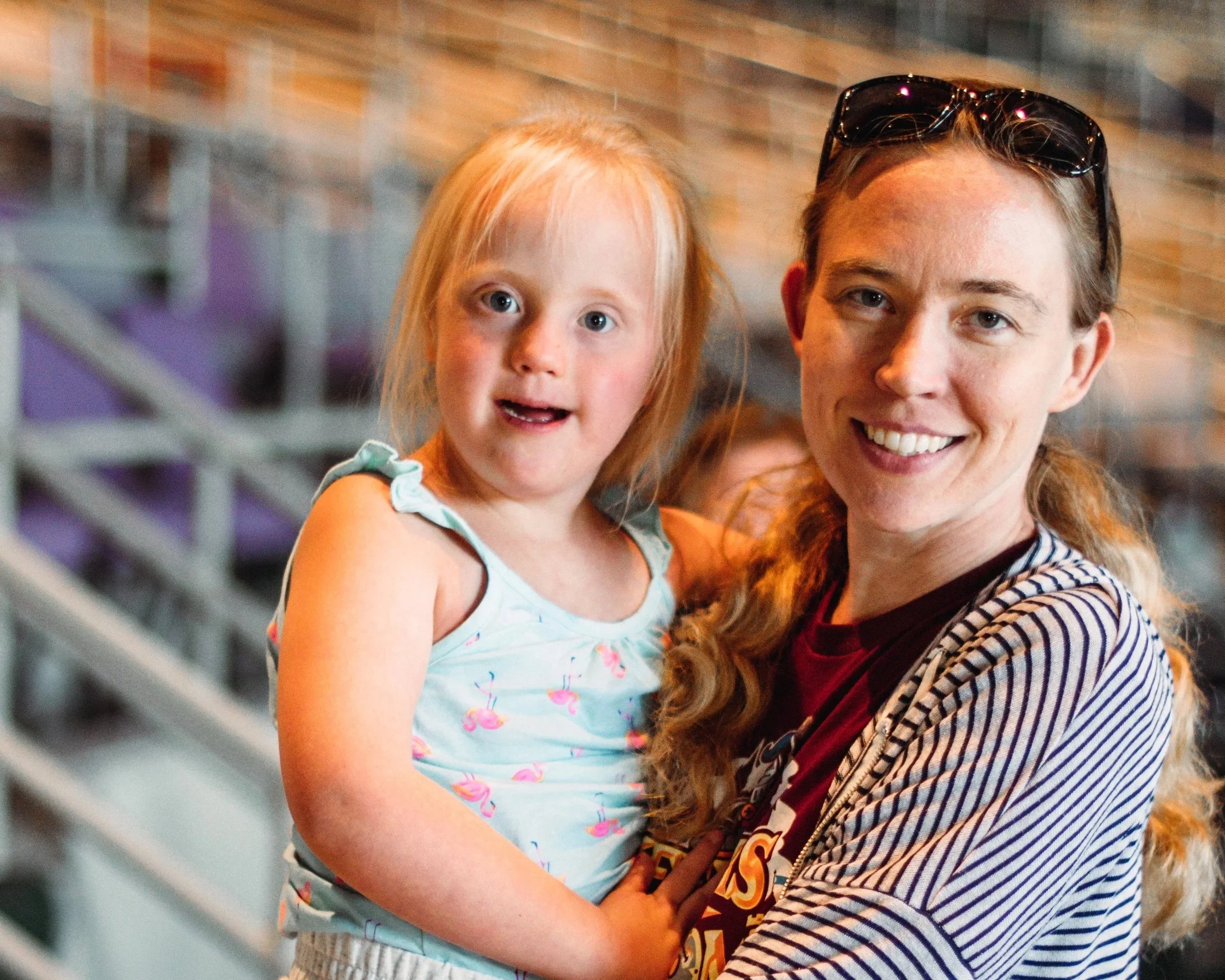 A smiling woman with long wavy hair holding a young girl with long red hair and bright blue eyes, against a background of a brick wall.