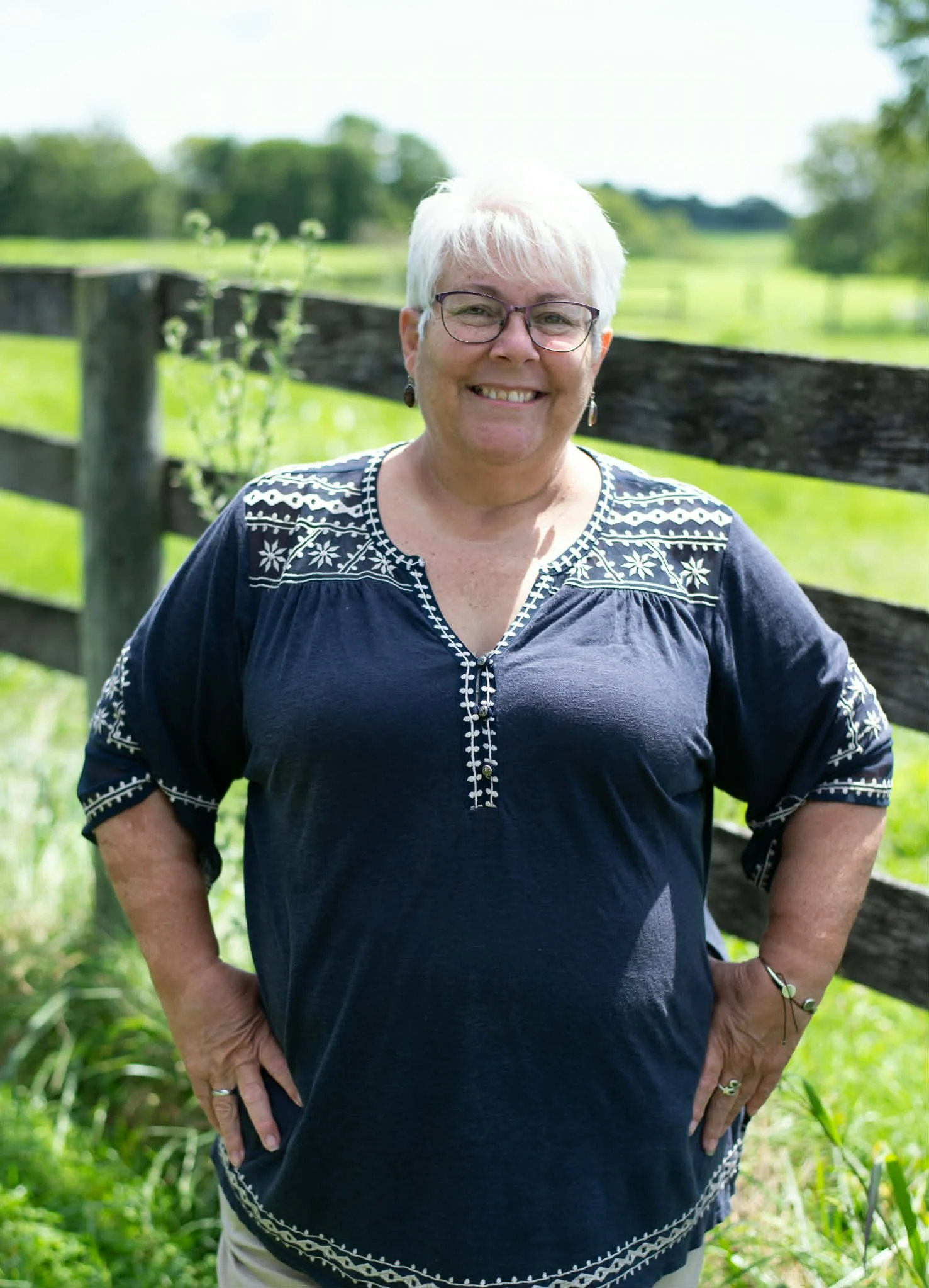 Close-up of a smiling woman with short gray hair, wearing glasses and a dark blue sleeveless top, standing against a bright blue background.