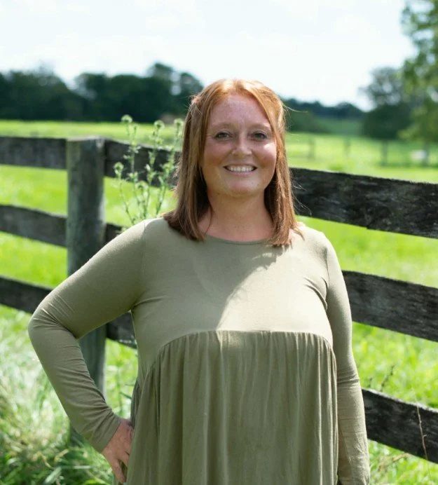 A smiling woman with red hair, freckles, and a cross necklace, wearing a light blue dress, standing in front of a blue textured background.