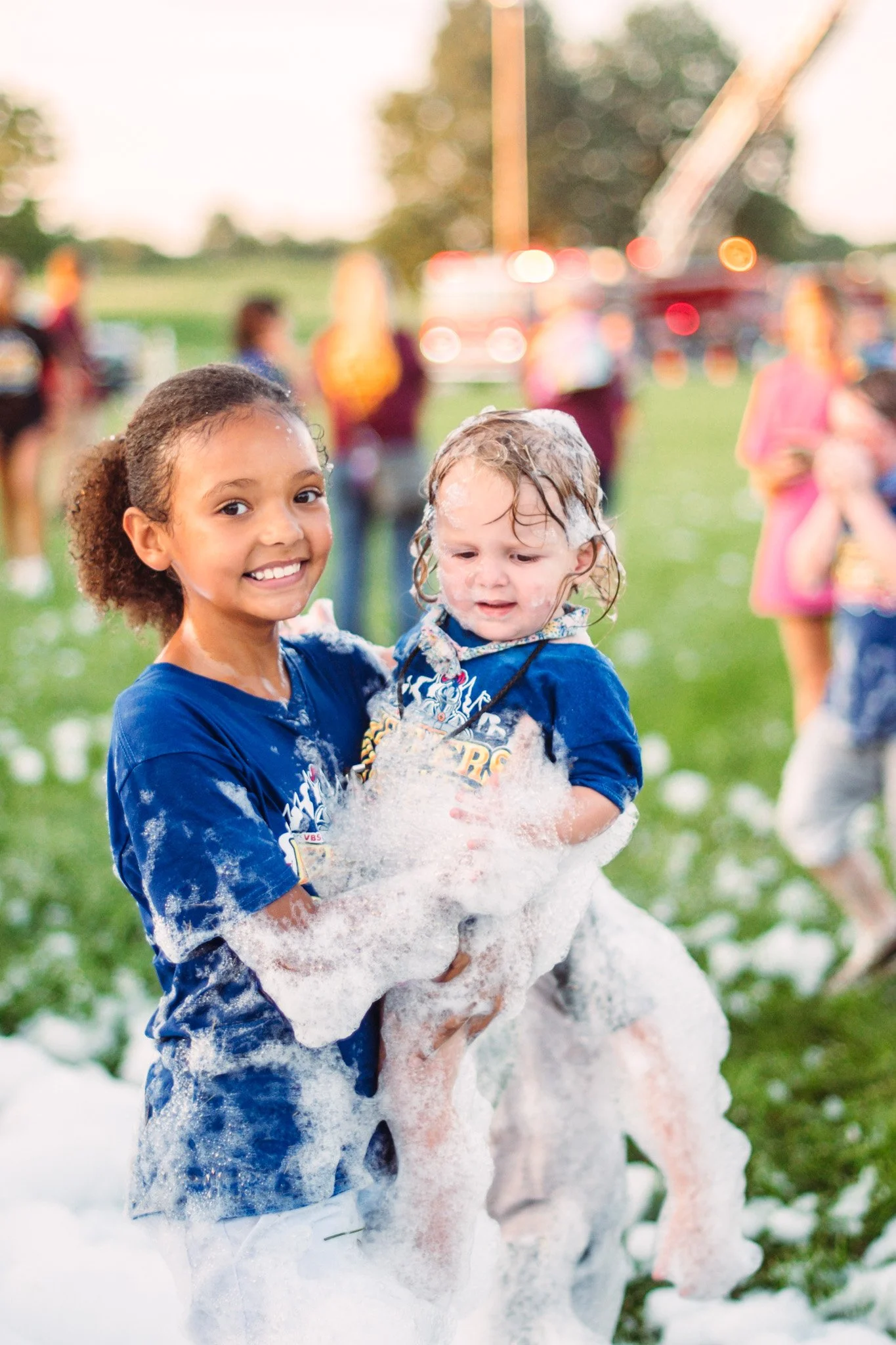 A young girl smiling and holding a smaller child covered in foam during a foam party outdoors, with other children and adults in the background.
