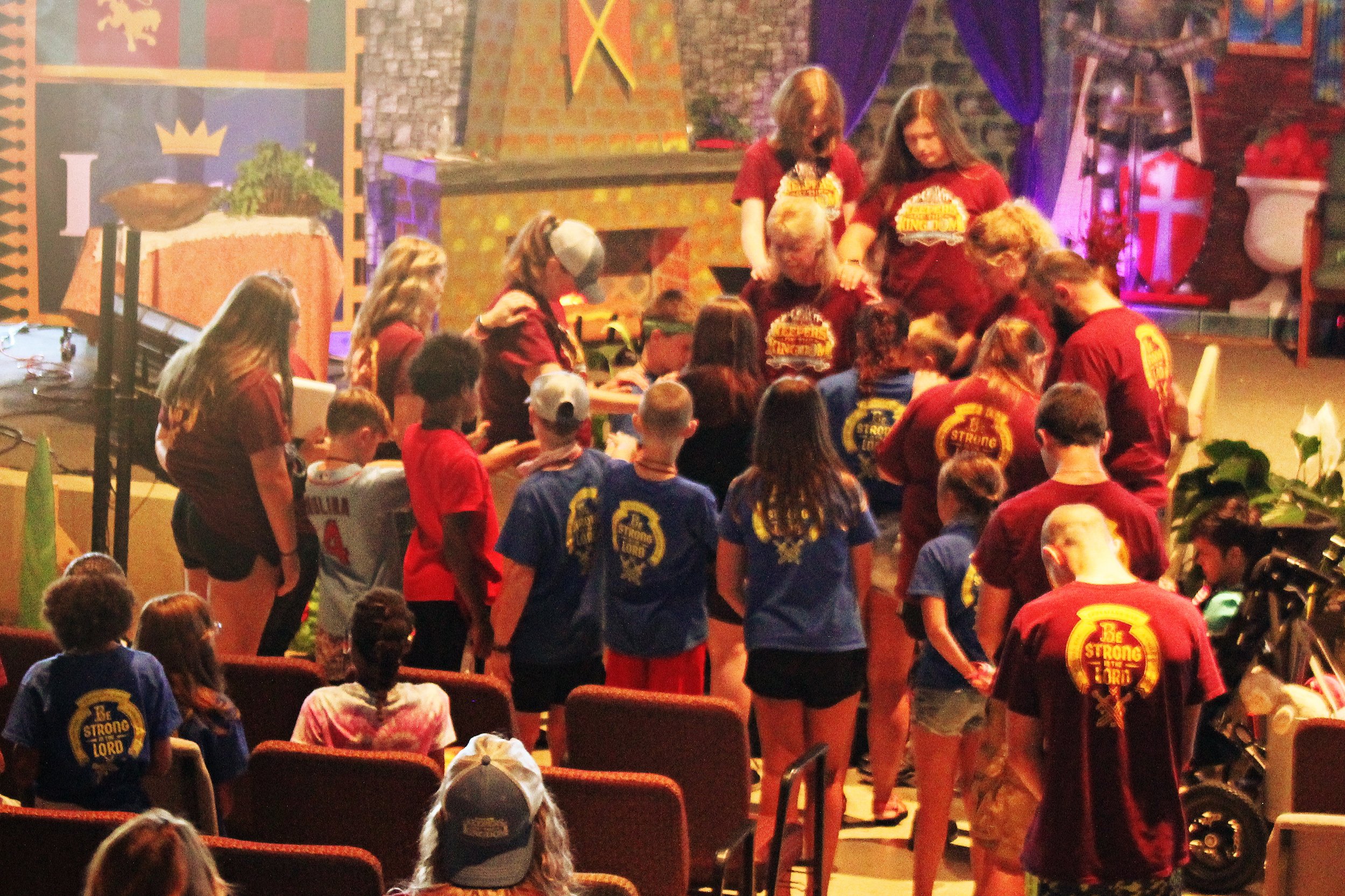 Group of young people gathered indoors, some wearing matching blue and red shirts with a 'Be Strong Lord' logo, participating in an event with stage decorations including a castle replica, banners, and colorful props.