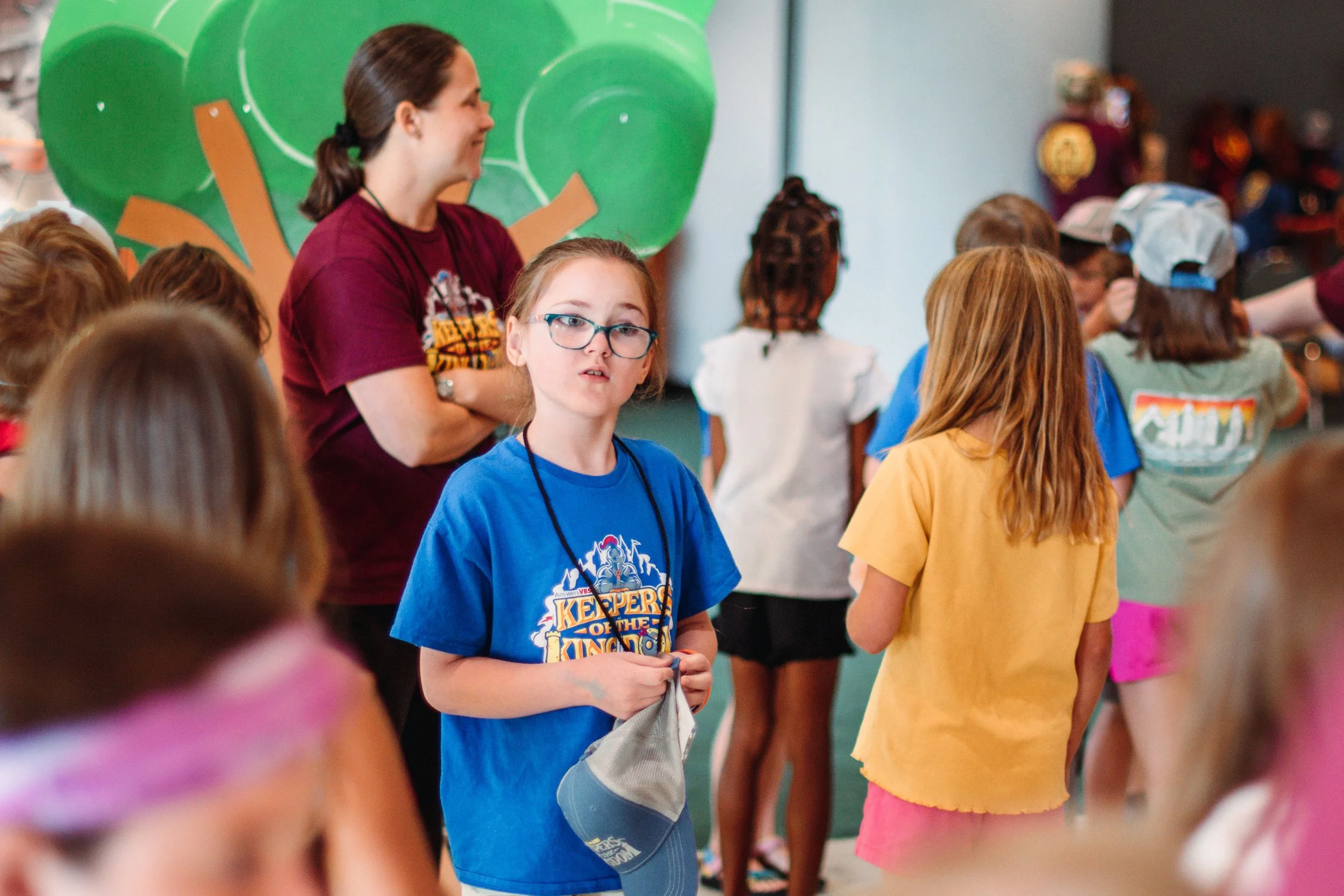 Children in a room, some wearing colorful shirts and hats, engaging in an activity. A woman with a ponytail and maroon shirt stands among them, smiling. The background features a large green and orange decorative element on the wall.