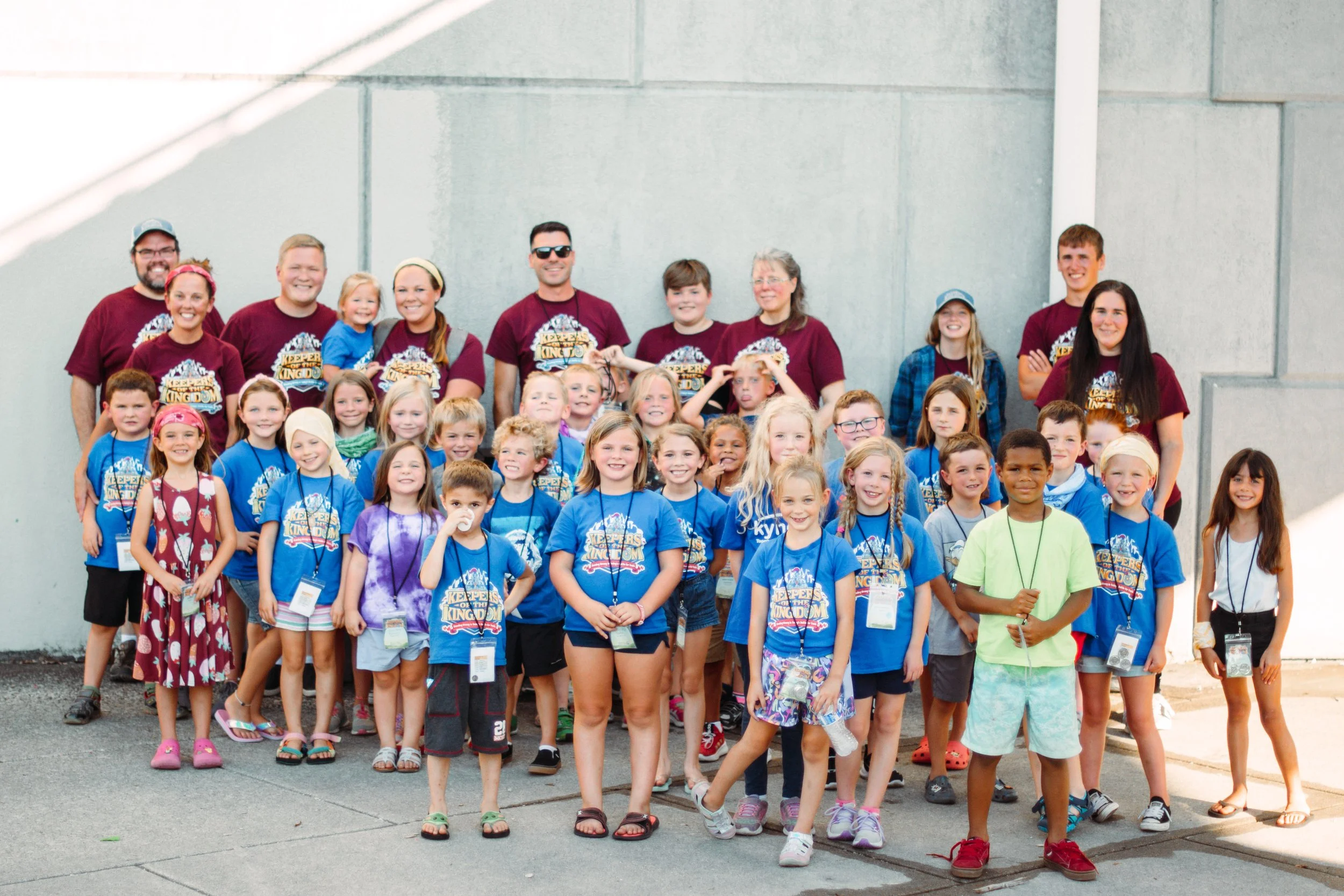 Group of children and adults posing for a photo outdoors against a plain wall, many wearing matching blue and maroon T-shirts with a logo, some with name tags, smiling and standing close together.