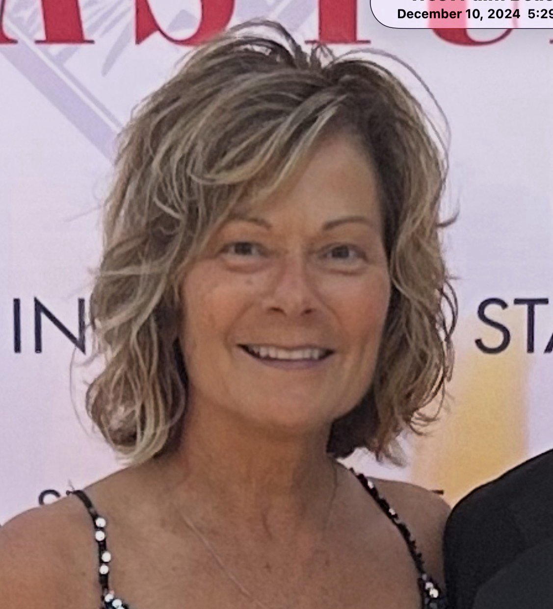 A woman with shoulder-length blonde hair, smiling, wearing a black sleeveless top and a cross necklace, standing against an orange background.