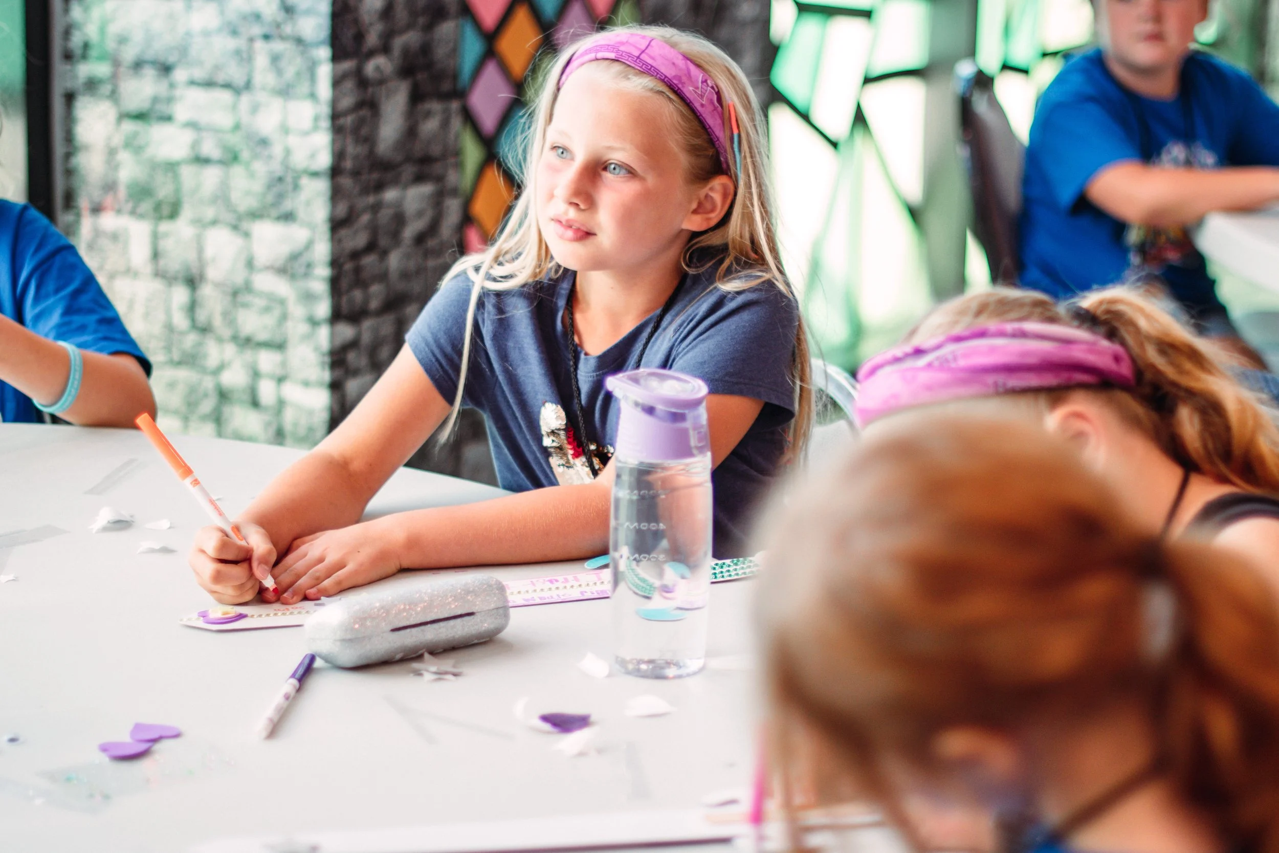 A young girl with blonde hair, wearing a purple headband, sitting at a table with paper, markers, and heart-shaped cutouts during an arts and crafts activity. She appears focused and is holding a red marker. In the background, there are other children and colorful geometric wall decorations.