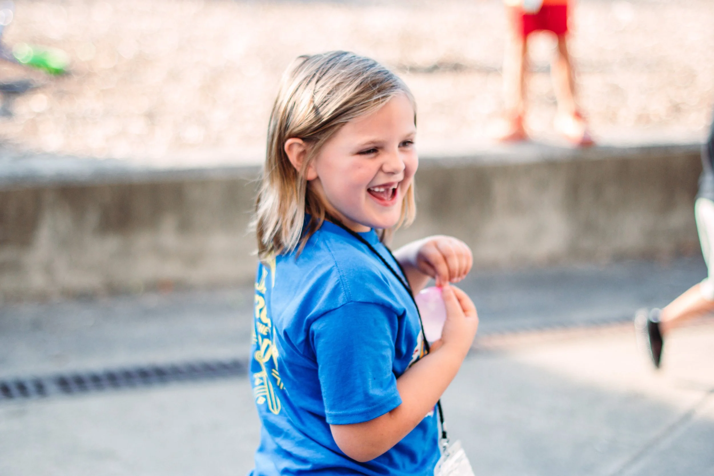 A young girl with blonde hair laughing and smiling, wearing a blue sports shirt, in an outdoor setting.