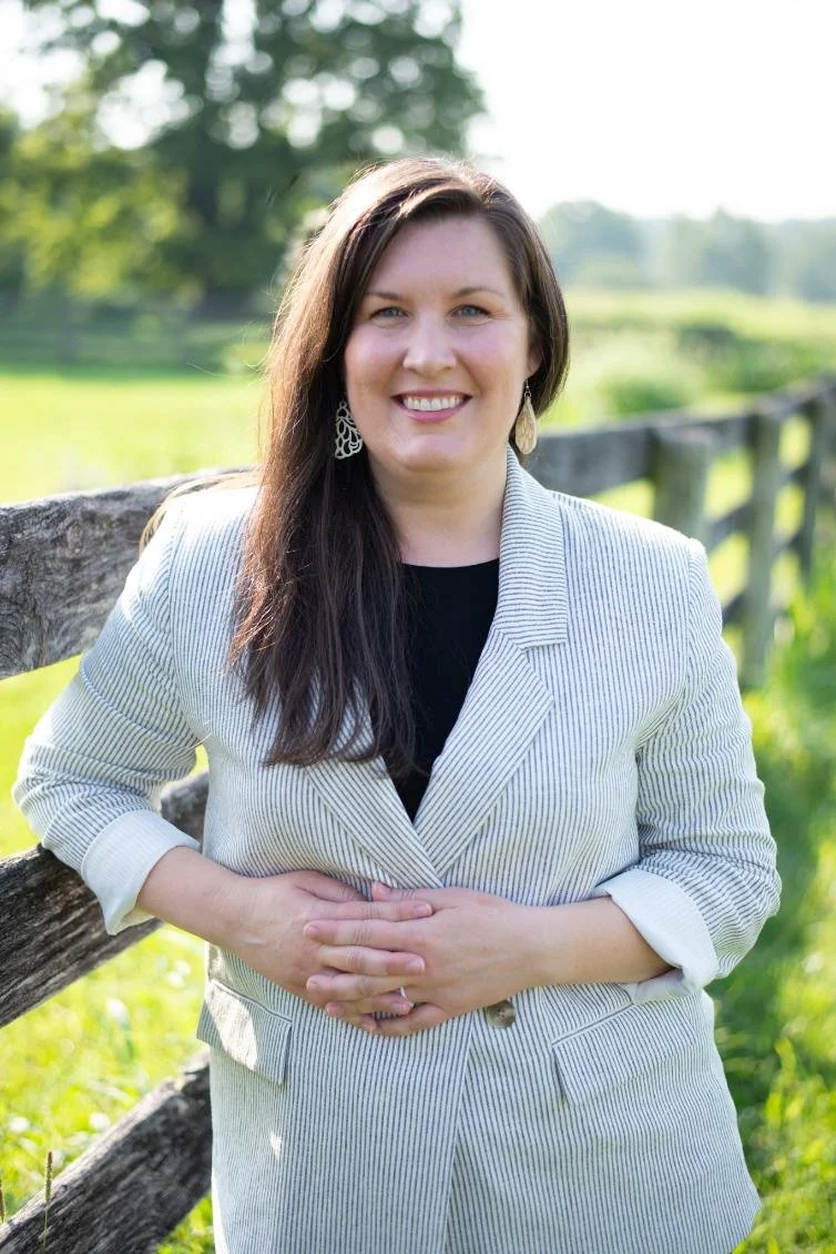 A smiling woman with long dark hair, wearing earrings, a navy blue top, a beige cardigan, and a gold necklace, standing against a bright blue background.