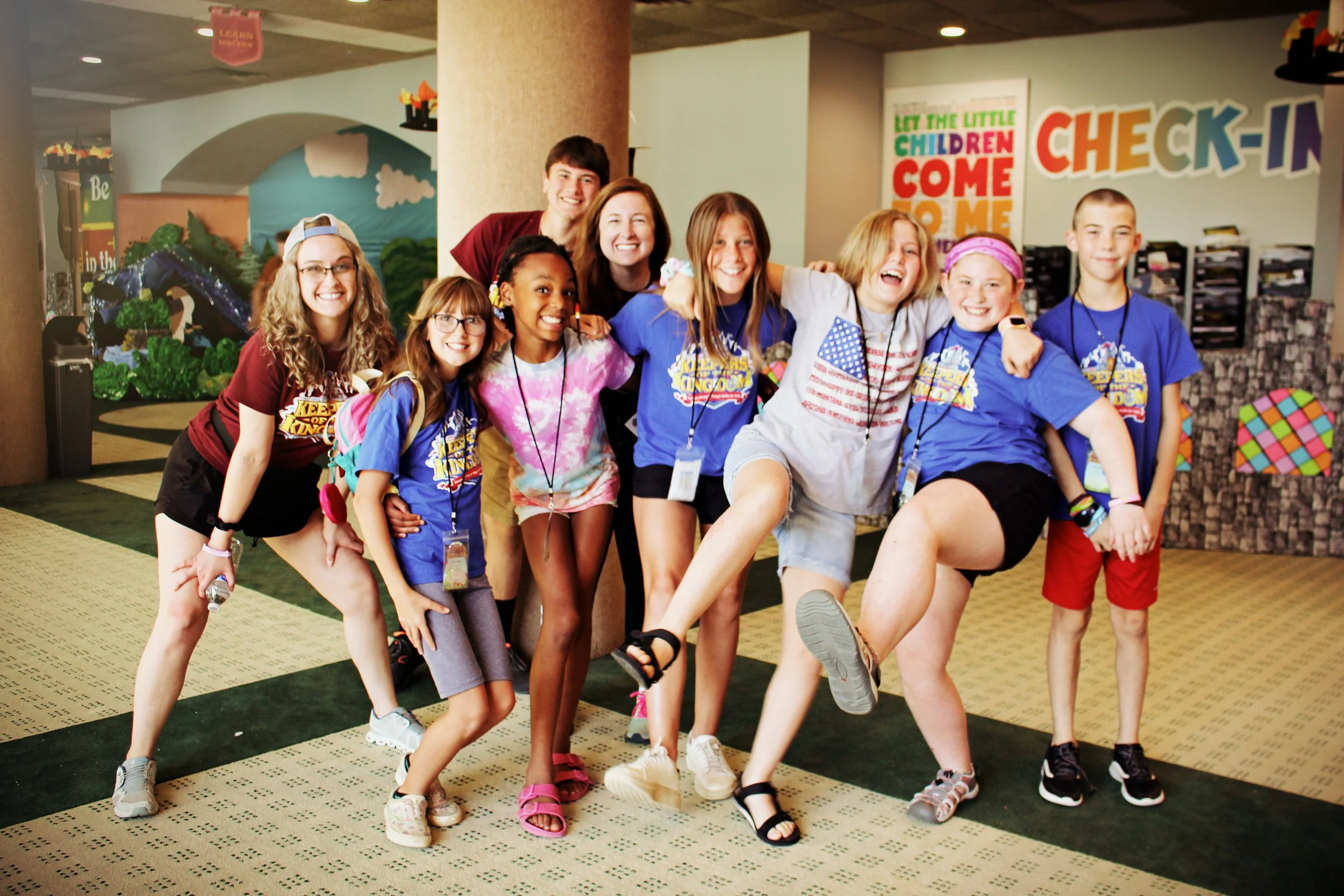 A group of children and two adults, smiling and posing together inside a colorful indoor facility, possibly a children's play area or community center, with various decorations and signs on the walls.