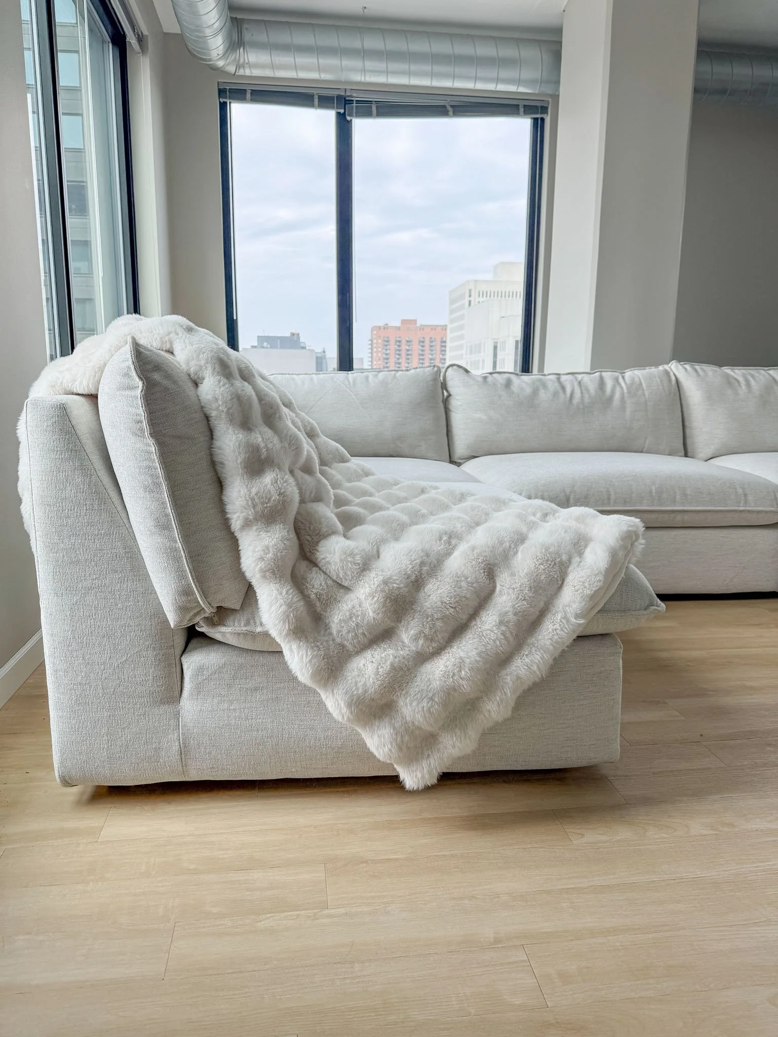 A cozy living room corner with a white sectional sofa draped with a plush faux fur throw blanket, large windows showing downtown Minneapolis and a cloudy sky outside.