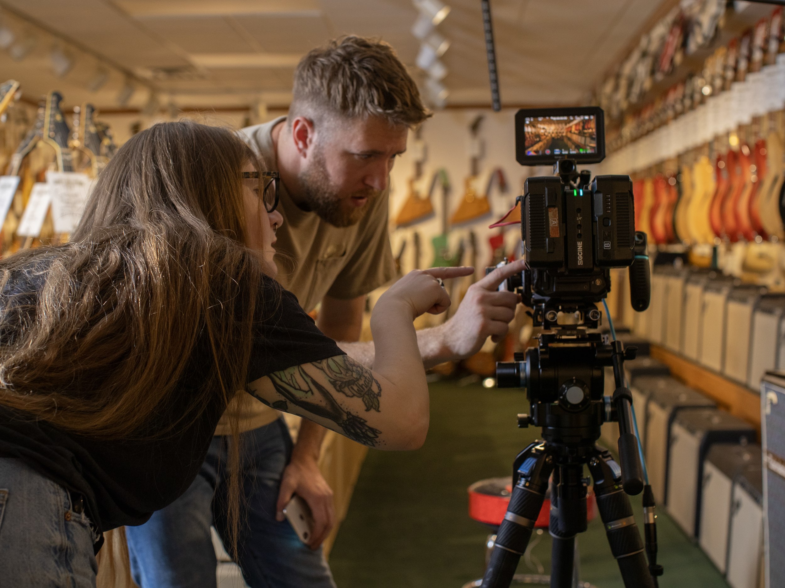Two people filming with a professional camera in a music store with guitars on the wall.
