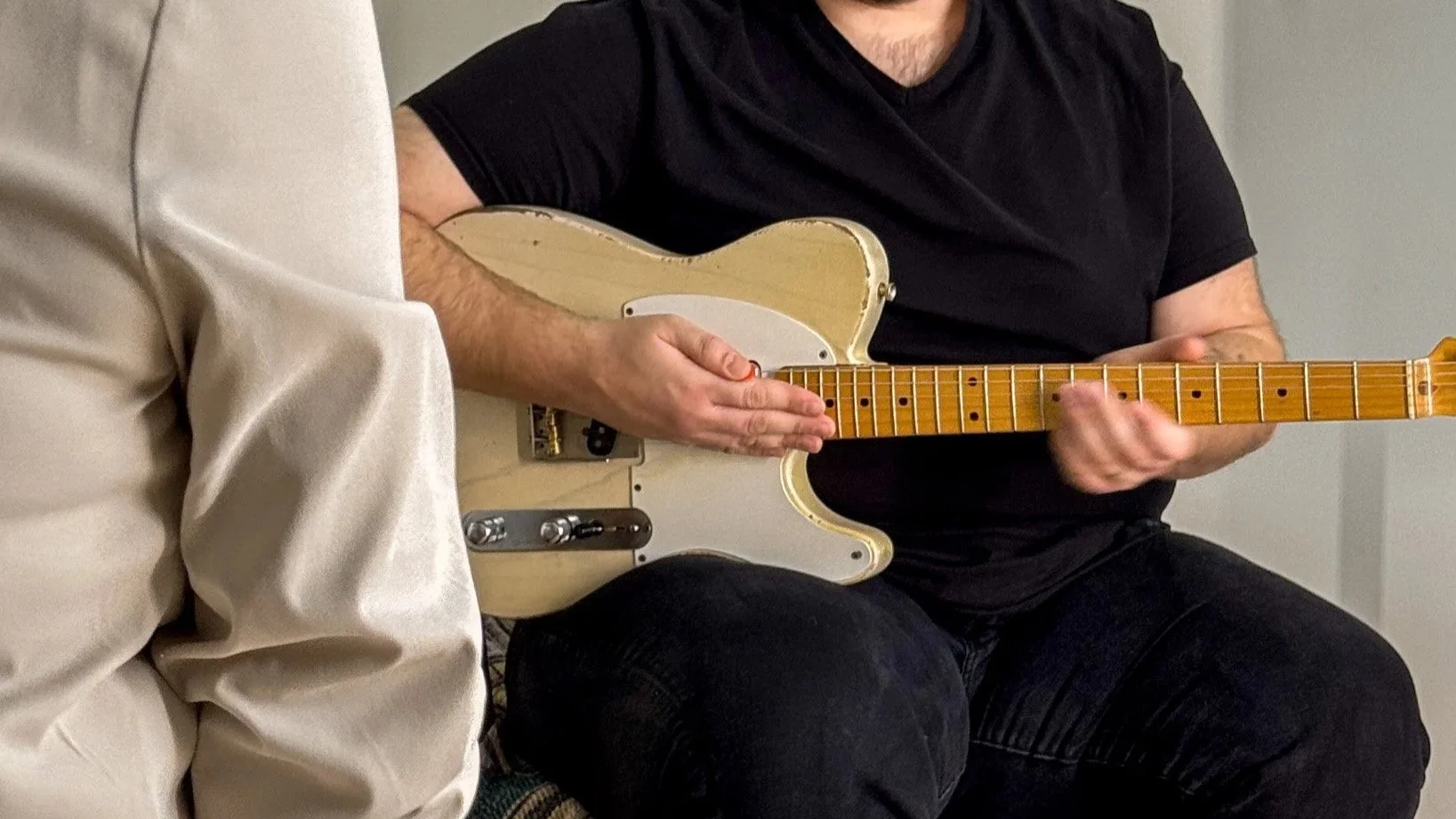 A person playing an electric guitar, wearing a black shirt, sitting with a white couch and another person in beige visible nearby.