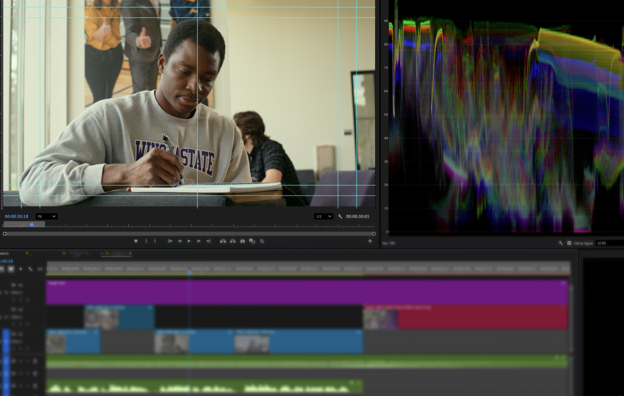 A young man wearing a gray sweatshirt with 'WISCONSIN' on it sits at a desk, signing a notebook. Behind him, two people are standing and talking in a room with a window and a small monitor. The image is part of a video editing software interface showing a timeline at the bottom and a color spectrum on the right.
