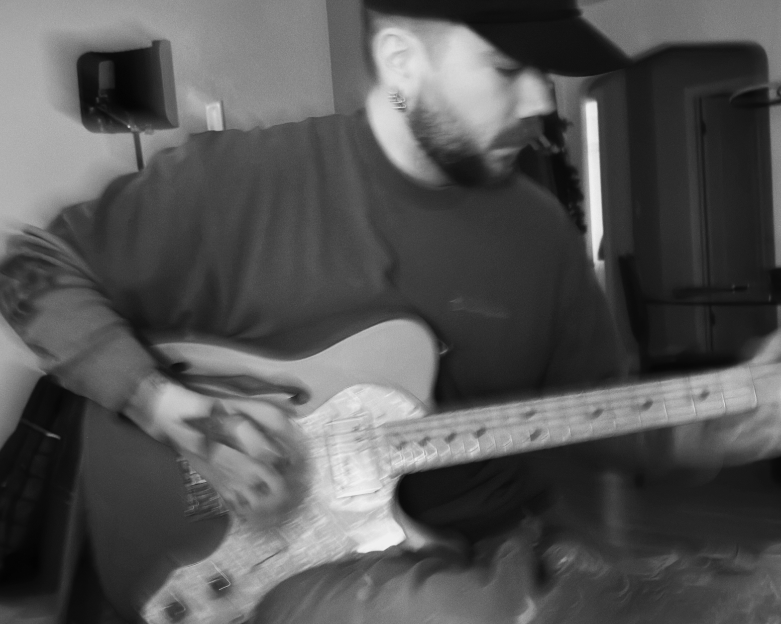 A man with a beard, wearing a black baseball cap and a dark long-sleeve shirt, playing an acoustic guitar in a room with a framed picture and a speaker on the wall.