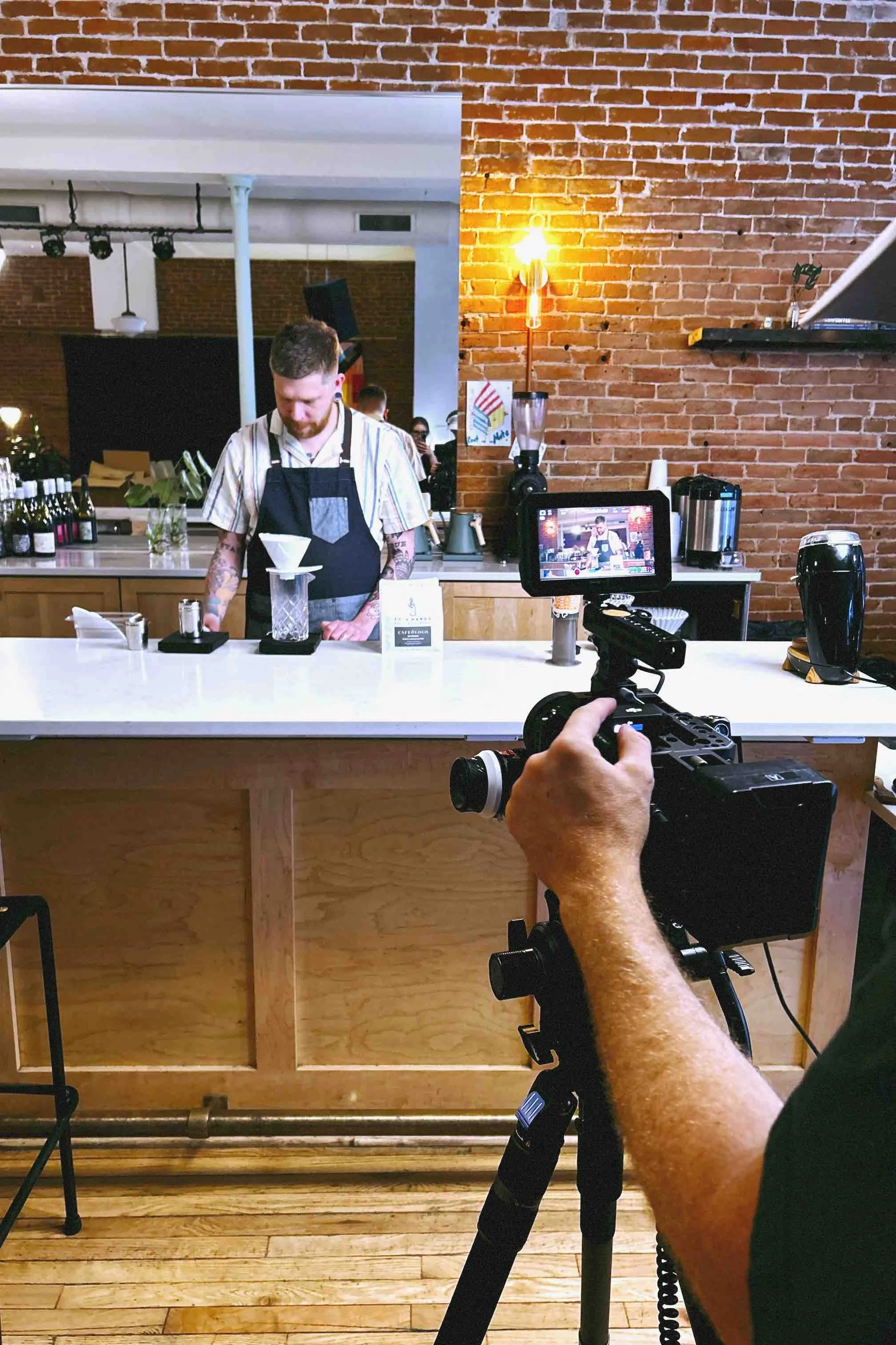 BS Creative's director of photography is filming a barista preparing a coffee behind a white counter in a coffee shop with an industrial brick wall background.