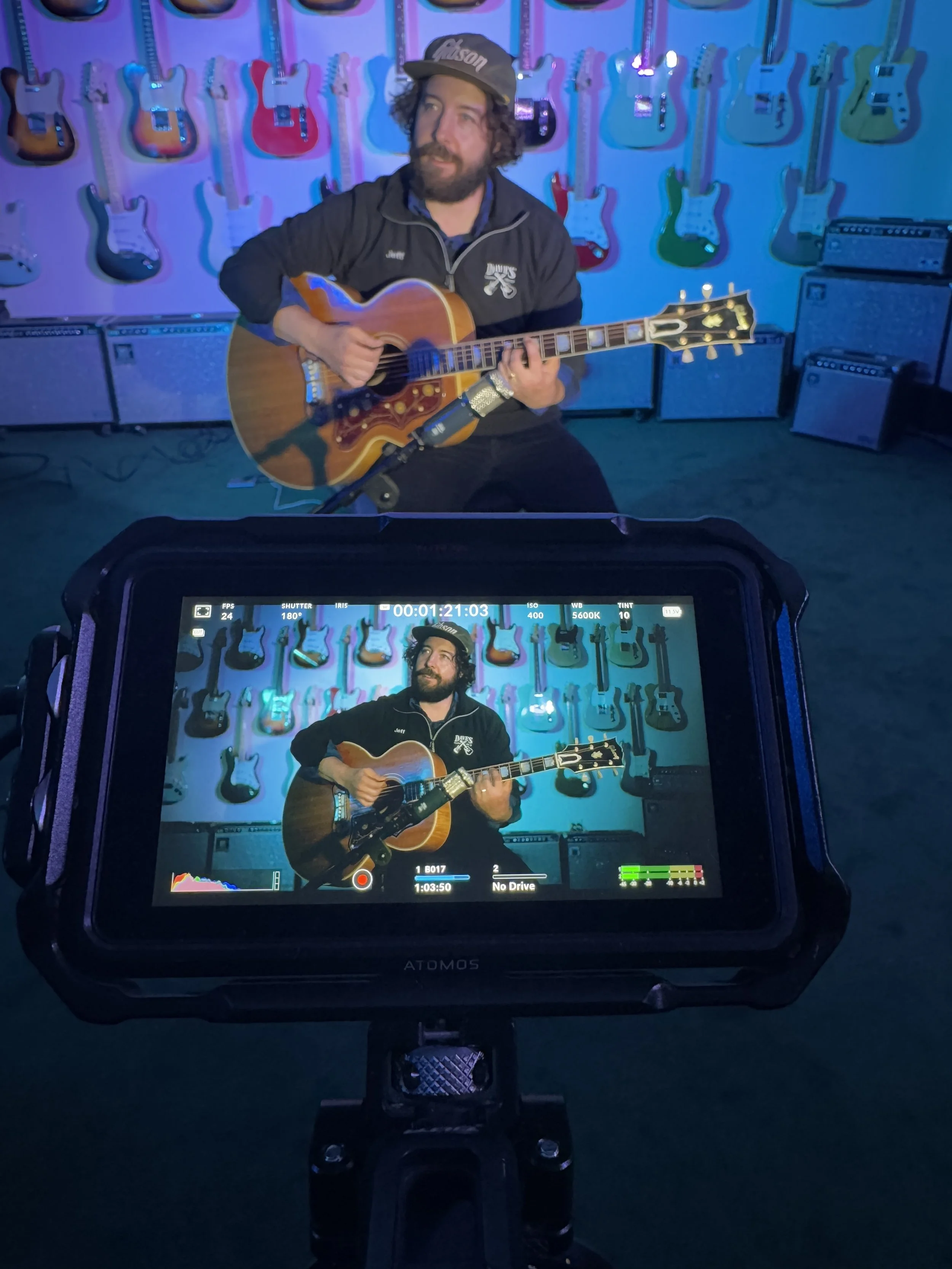 Man with curly hair and beard wearing a cap and black jacket playing acoustic guitar in front of a wall of colorful electric guitars, with a camera screen showing the same scene.