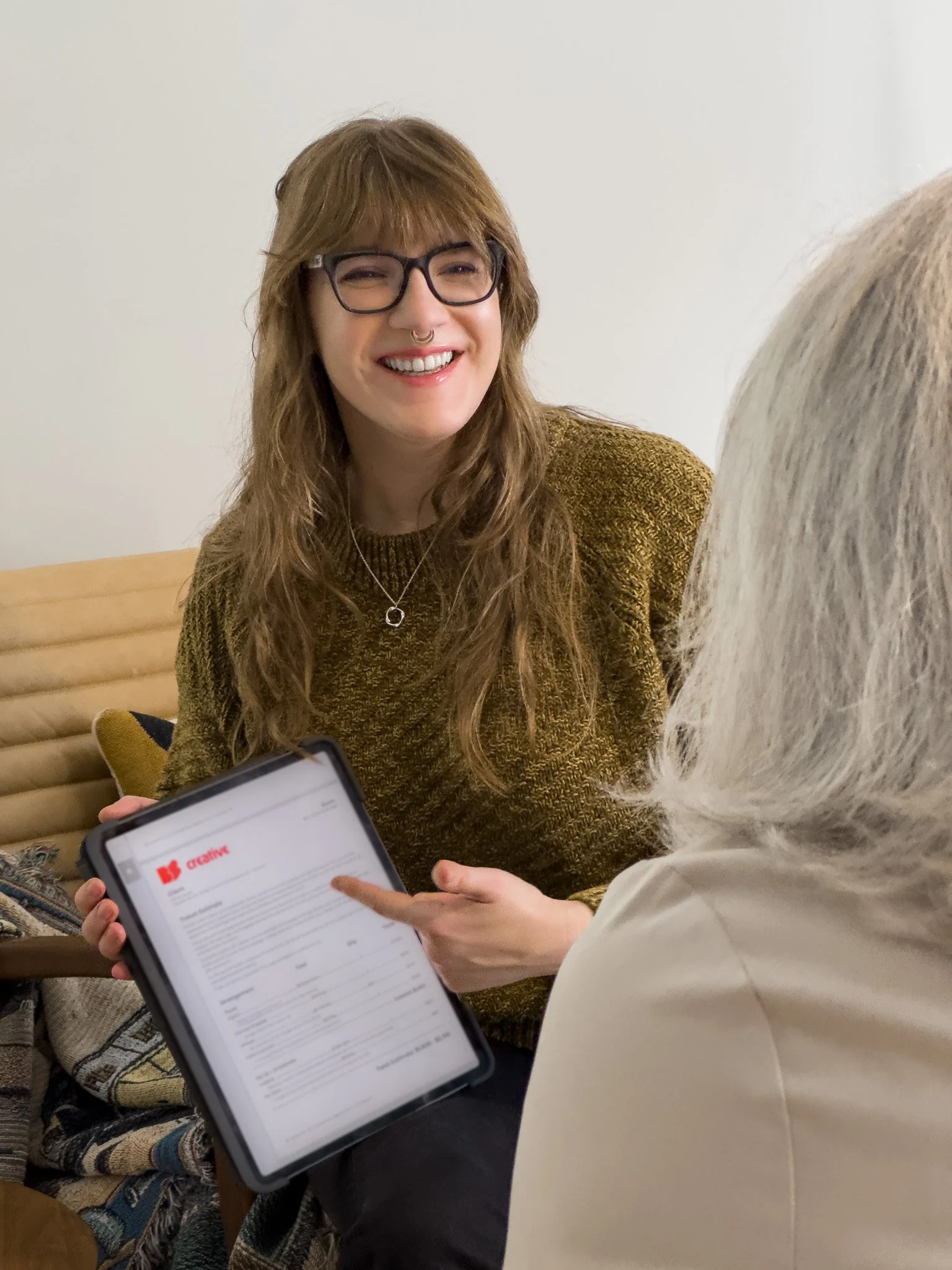 A young woman with long, wavy brown hair, glasses, and a septum piercing smiling while looking at an older person holding a tablet.
