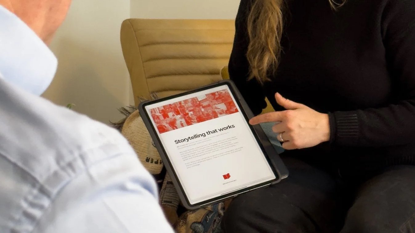 Two people sitting on a couch, one holding a tablet. The tablet screen shows a webpage with the title 'Storytelling that works' and a red graphic at the top.