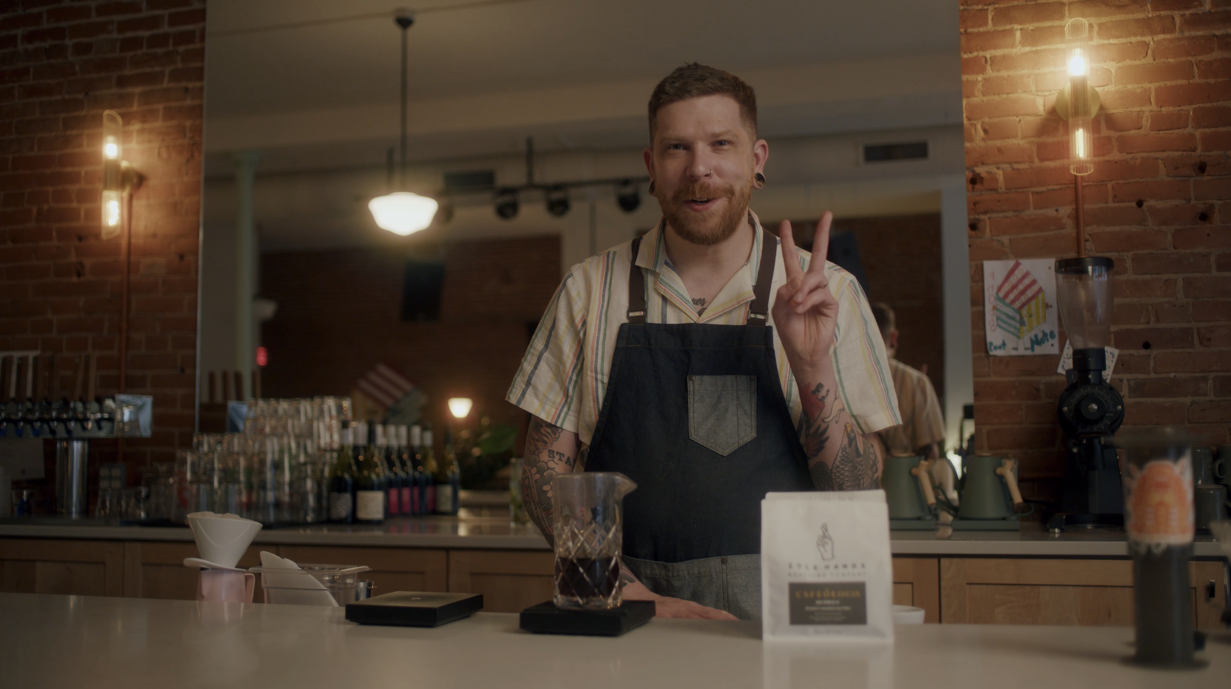 Root Note owner with tattoos and earrings wearing a striped shirt and apron, smiling and making a peace sign in a coffee shop with a brick wall and warm lighting.