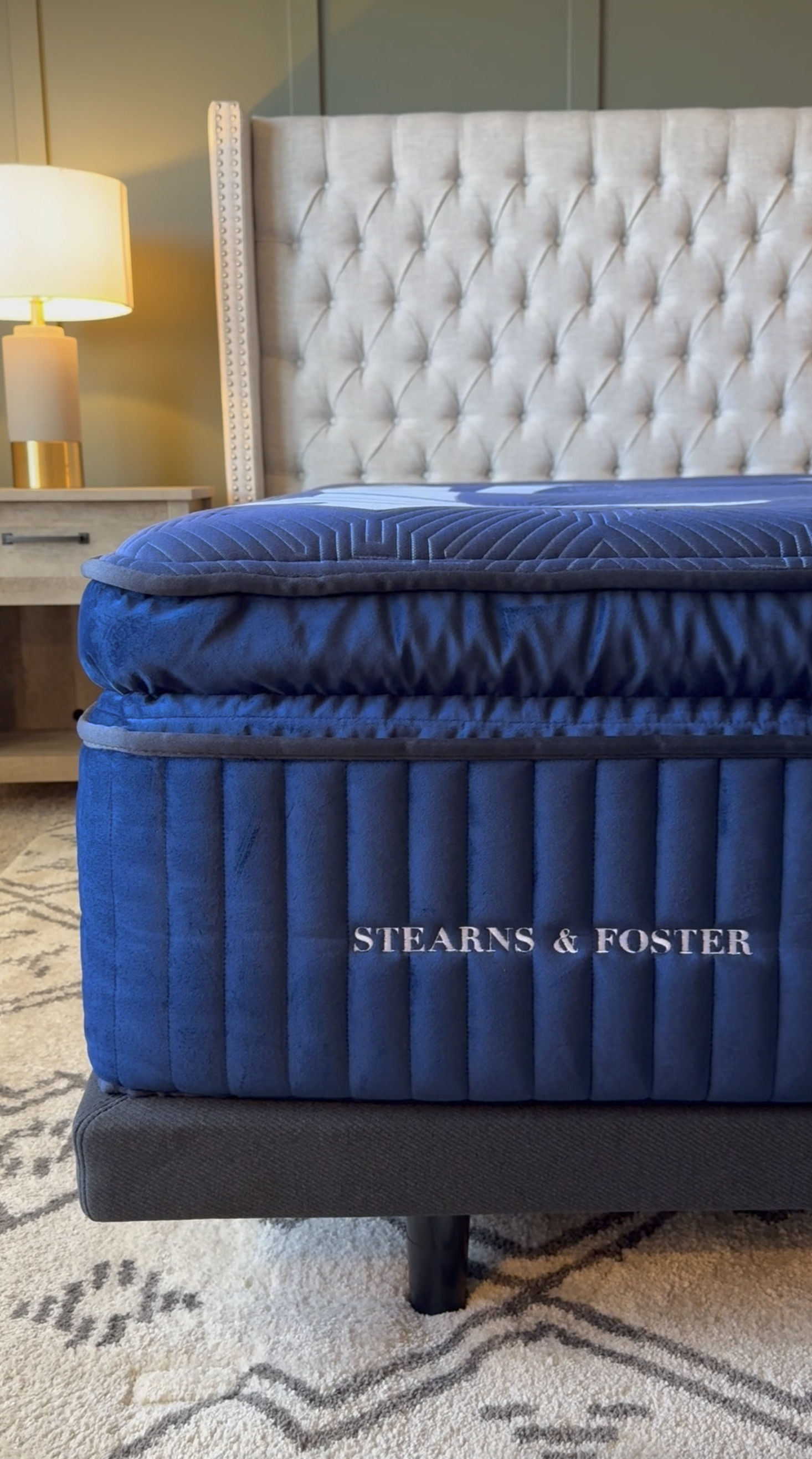 Close-up of a blue mattress with 'Stearns & Foster' embroidery, part of a bed in a bedroom with a beige tufted headboard, a nightstand, and a lamp.