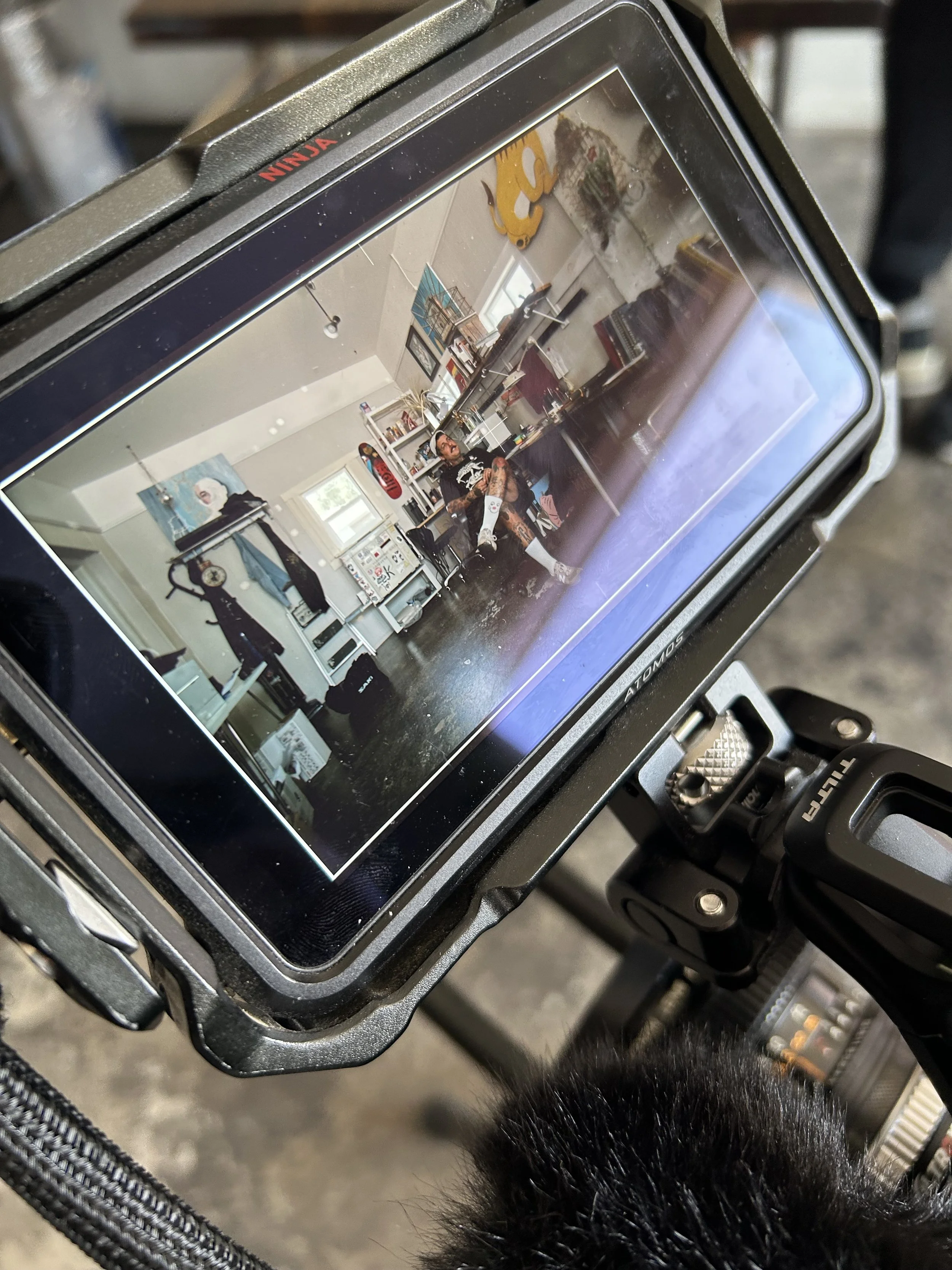 View of a person sitting in an art studio or workspace, surrounded by paintings, artwork, and furniture, seen through a camera monitor.