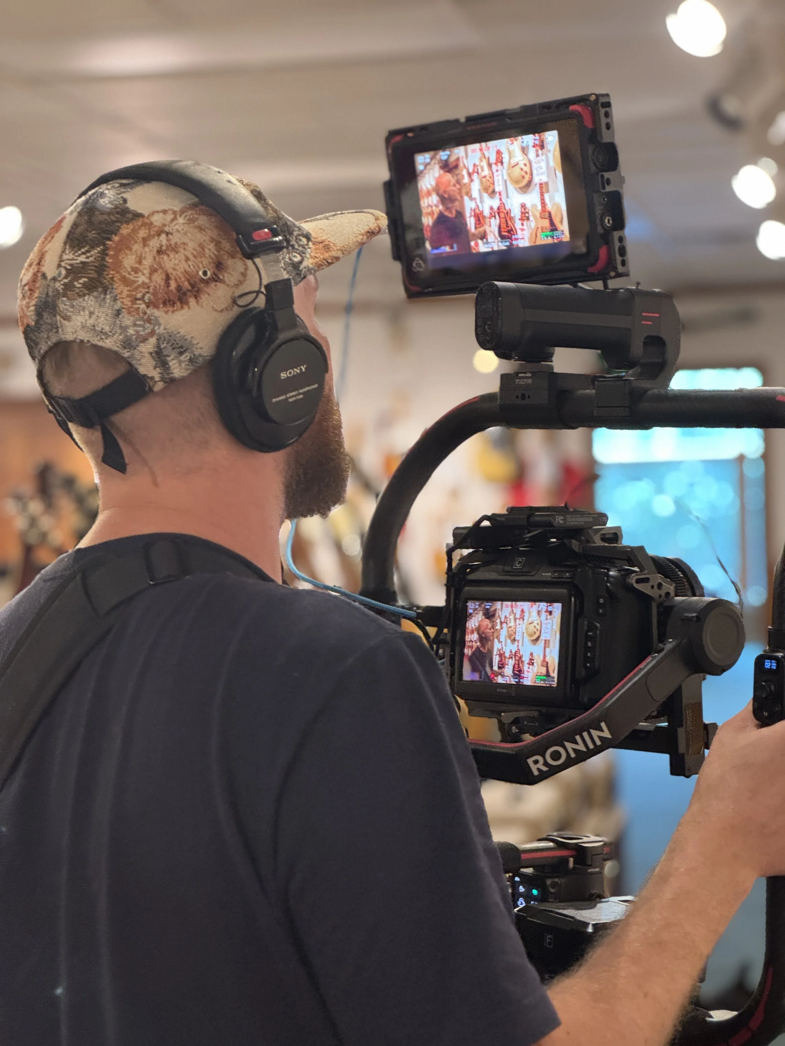 A man wearing a floral baseball cap and Sony headphones operating a professional camera on a stabilizer rig, filming an indoor scene with colorful decorations and a window in the background.