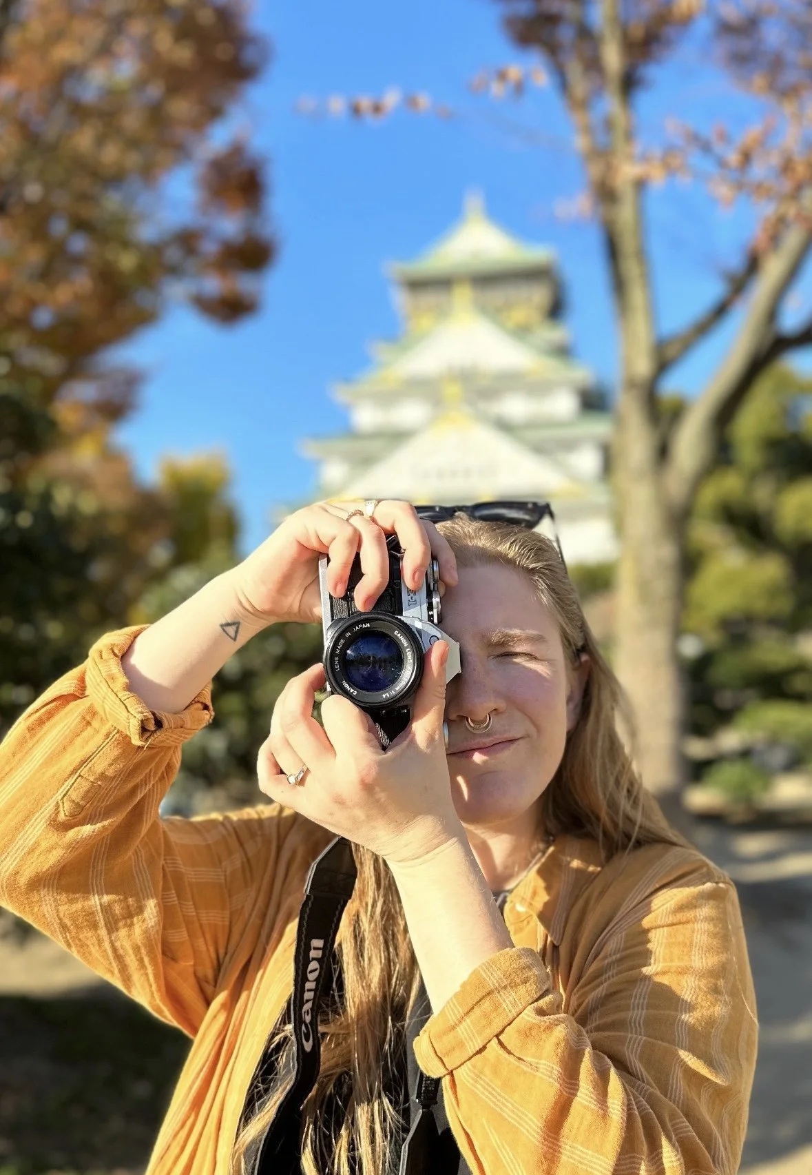 BS Creative co-founder, Sara Sampey, holds a camera outdoors, with Osaka Castle and trees in the background on a sunny day.