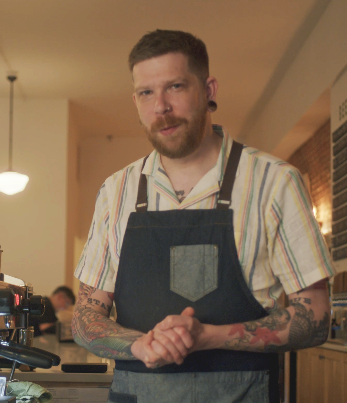 Coffee shop owner with a beard and tattoos, wearing a striped shirt and black apron, stands behind a counter in a café, looking at the camera.