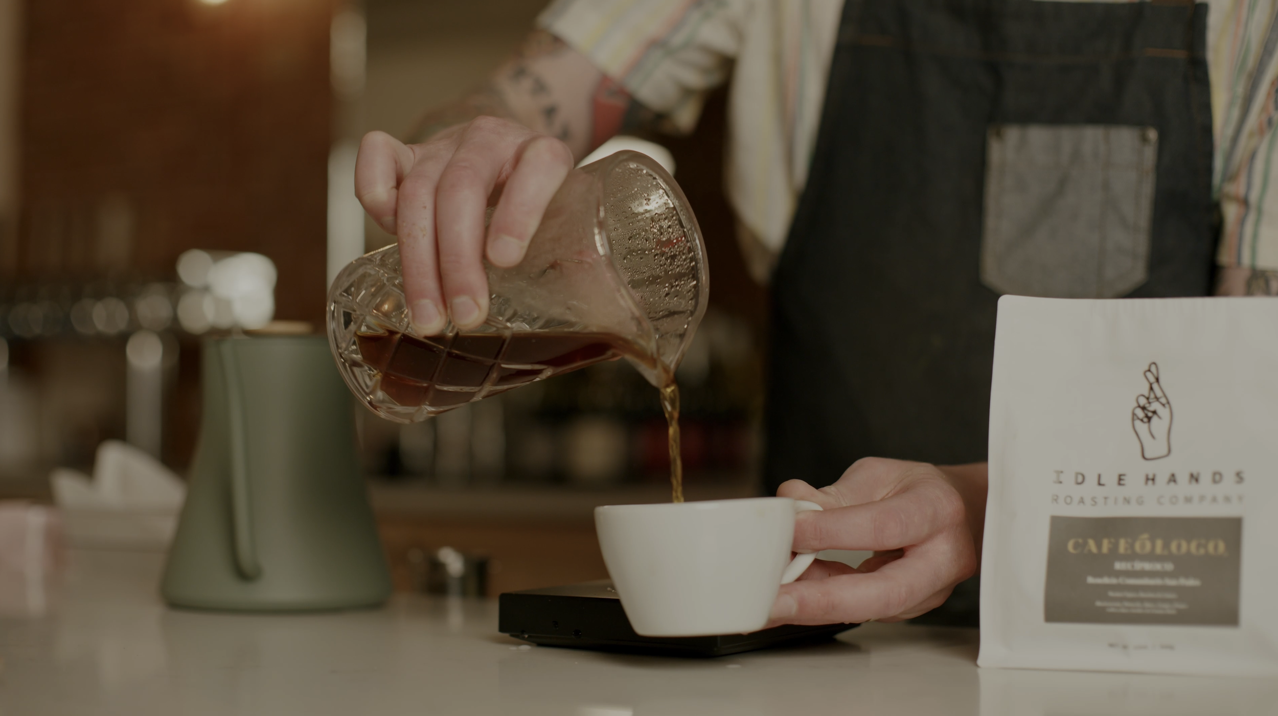 Barista pouring hot coffee from a glass pot into a white cup on a scale, with a bag of coffee labeled Idle Hands Roasting Company Cafe Logo in the foreground.