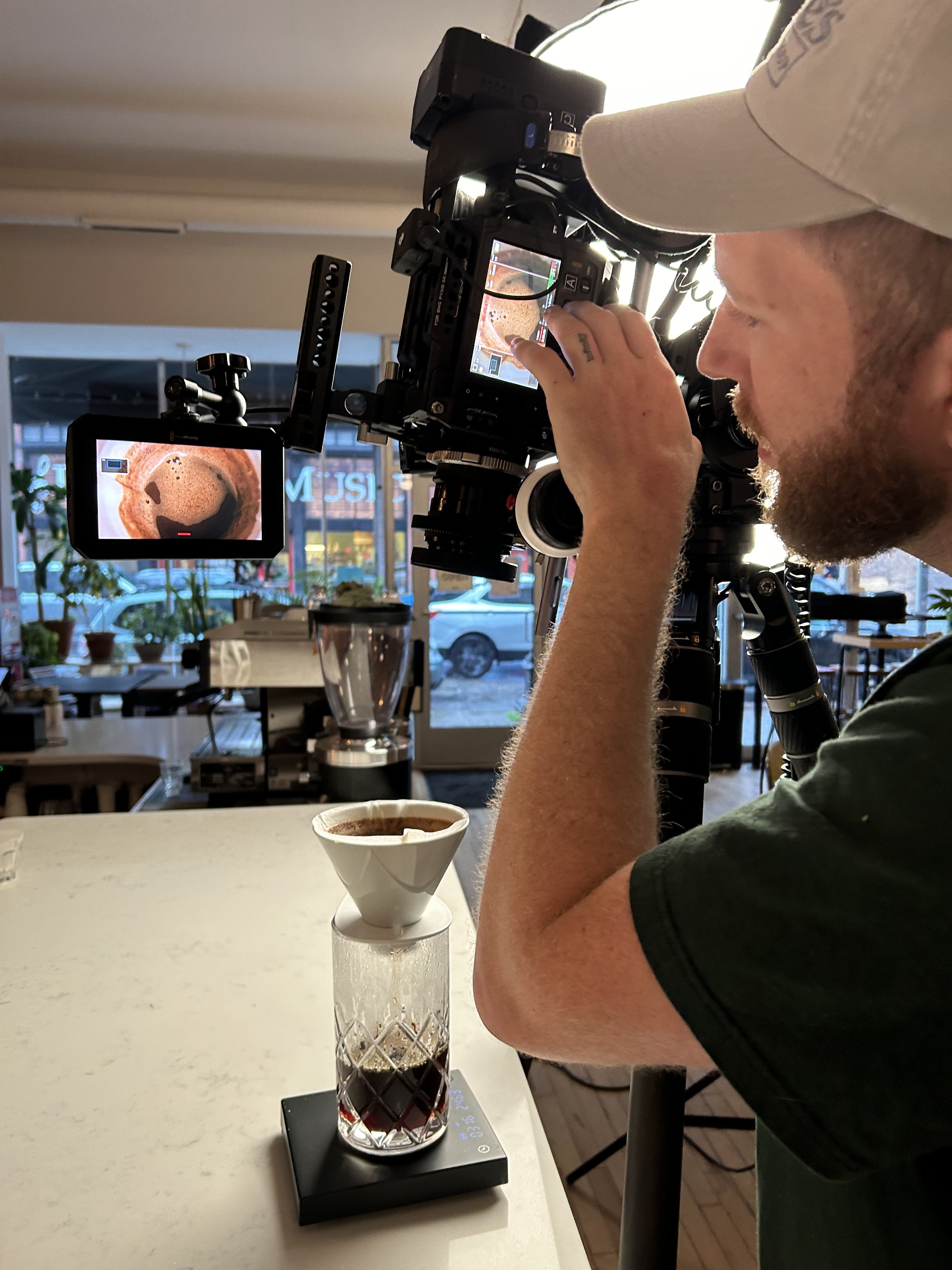 Videographer filming a coffee brewing process with a professional camera inside a cafe. The camera is focused on a glass of blooming coffee with a pour-over filter on top for a craft coffee brewing series.