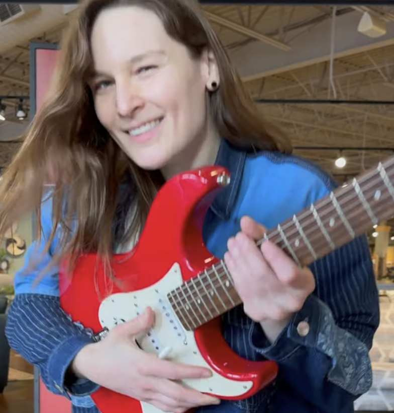 A woman with long brown hair smiling while holding a red electric guitar, in an indoor setting with a wooden ceiling.