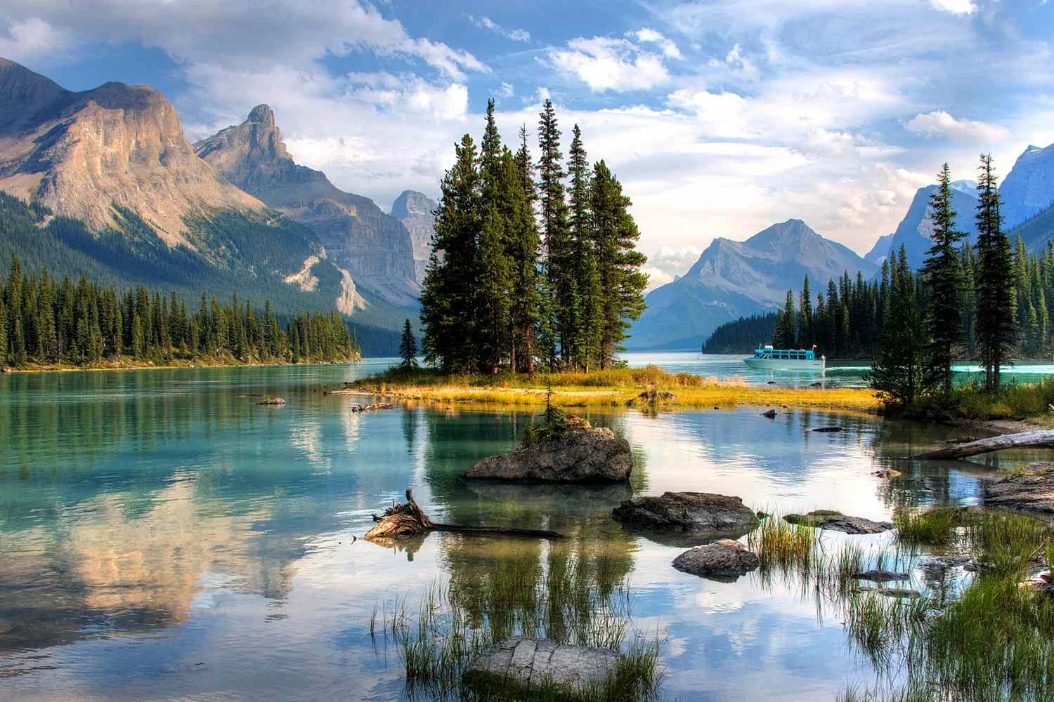 A scenic view of a large turquoise lake surrounded by tall pine trees and snow-capped mountains under a partly cloudy sky.