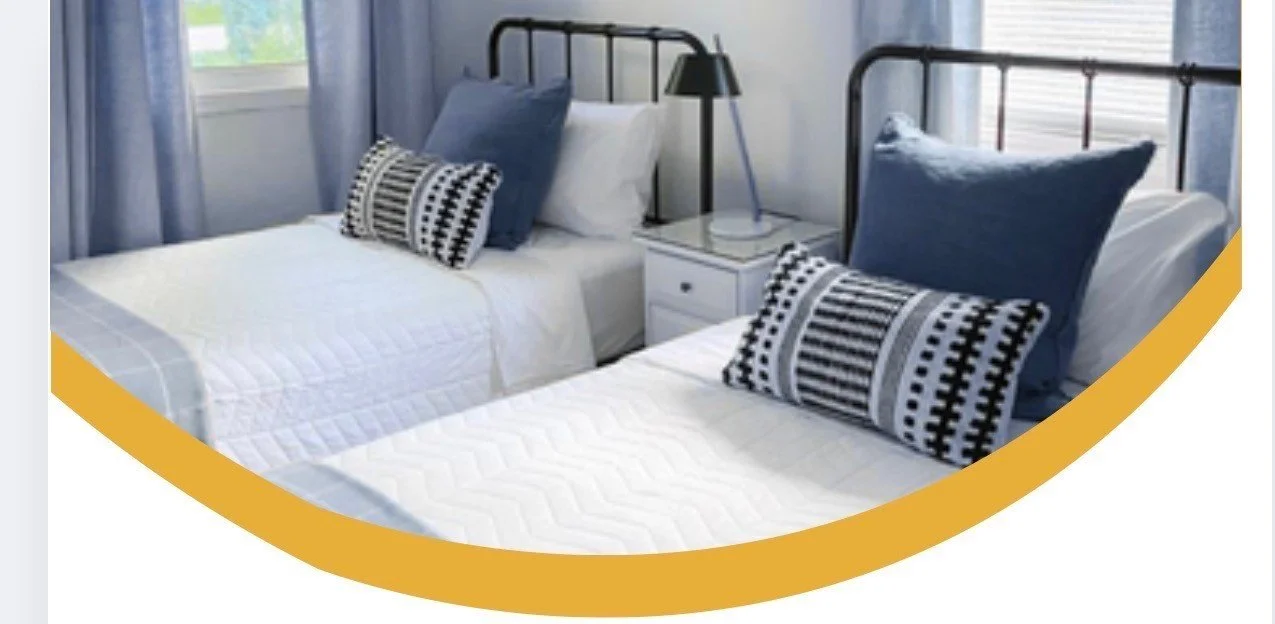 A bedroom with two twin beds, each with white quilted bedding, blue pillows, and black and white patterned throw pillows. There are black metal bed frames and a white nightstand between the beds with a lamp and a window with light blue curtains in the background.