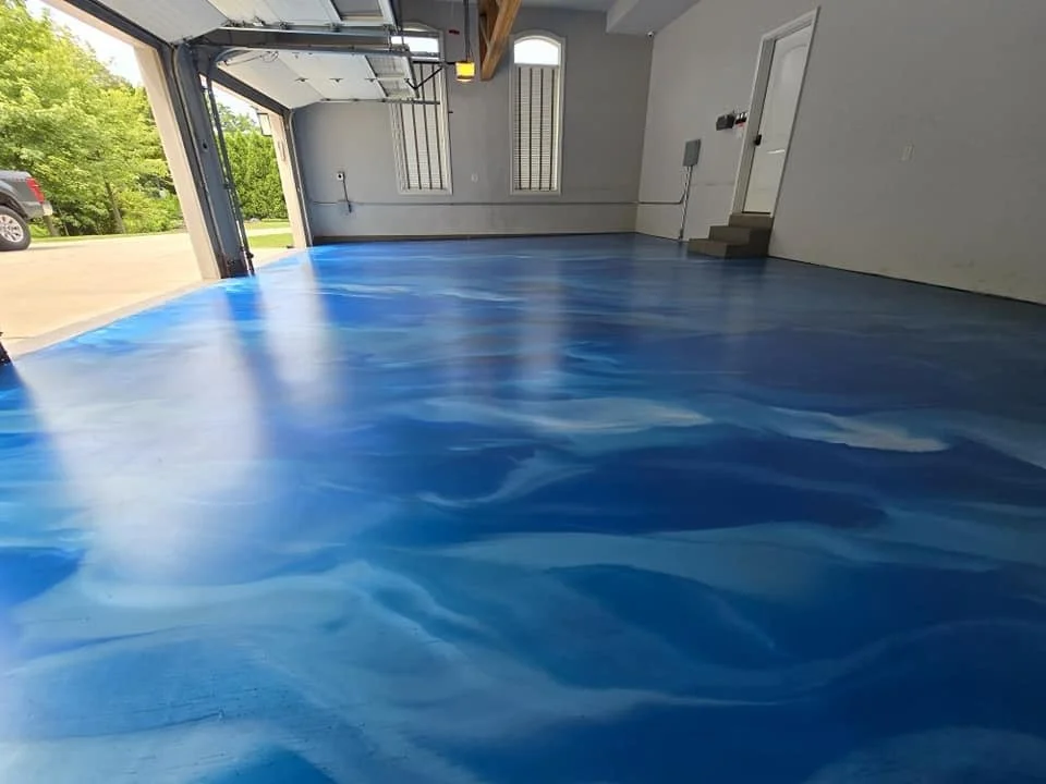 Interior of an empty garage with a blue, marbled epoxy floor, white walls, and large open garage door showing outside greenery and a parked vehicle.
