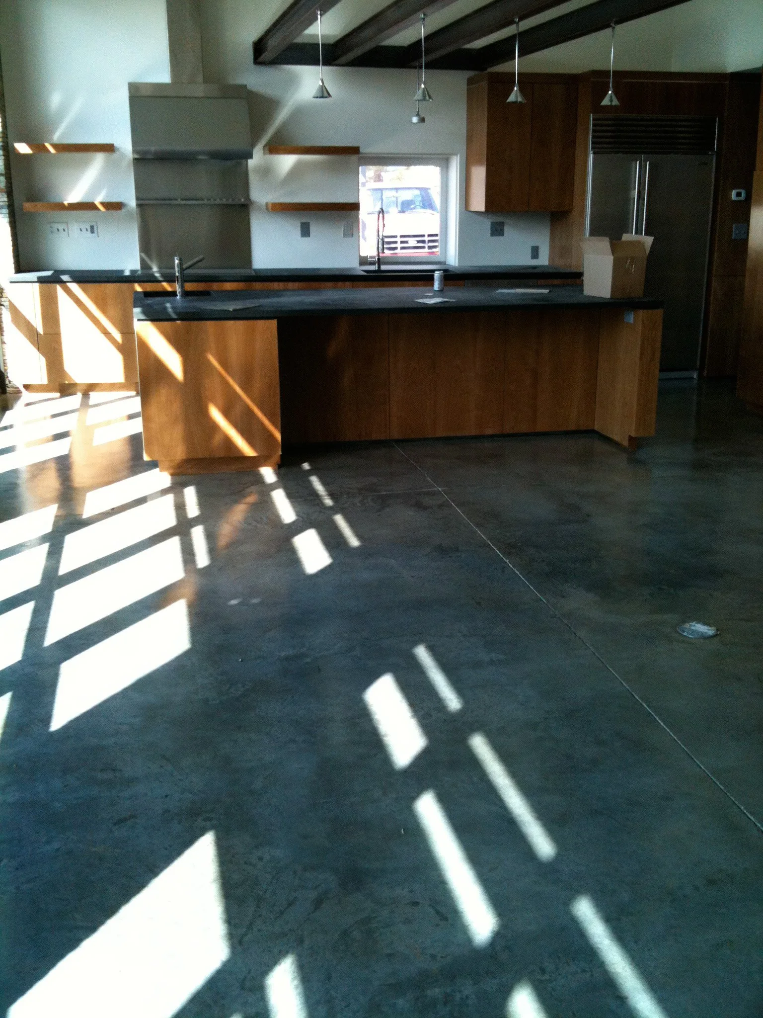 Sunlight creates shadow patterns on a modern kitchen with wooden cabinets, black countertops, and a concrete floor.