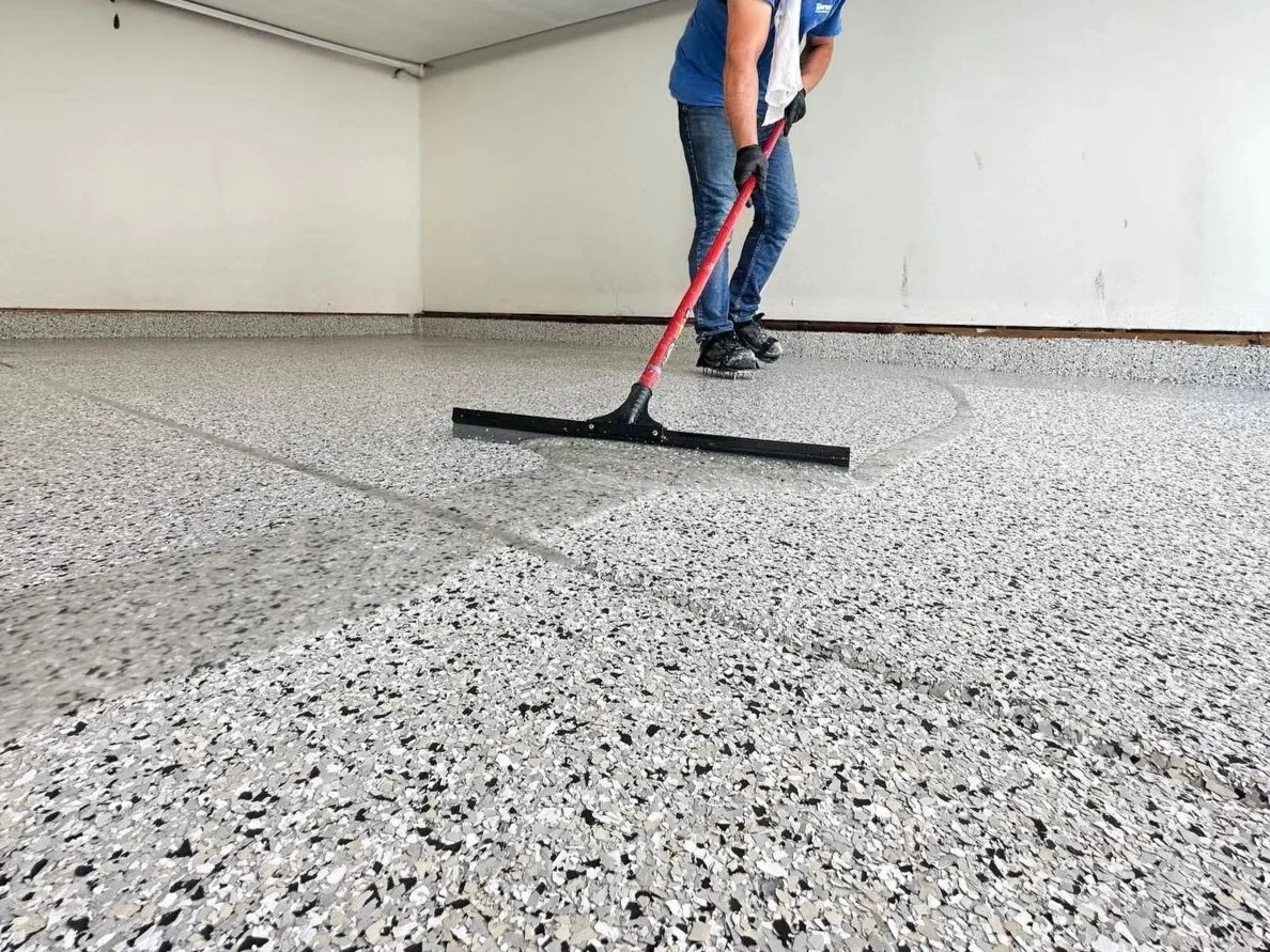 Person applying decorative concrete overlay with a squeegee in a room with terrazzo flooring.