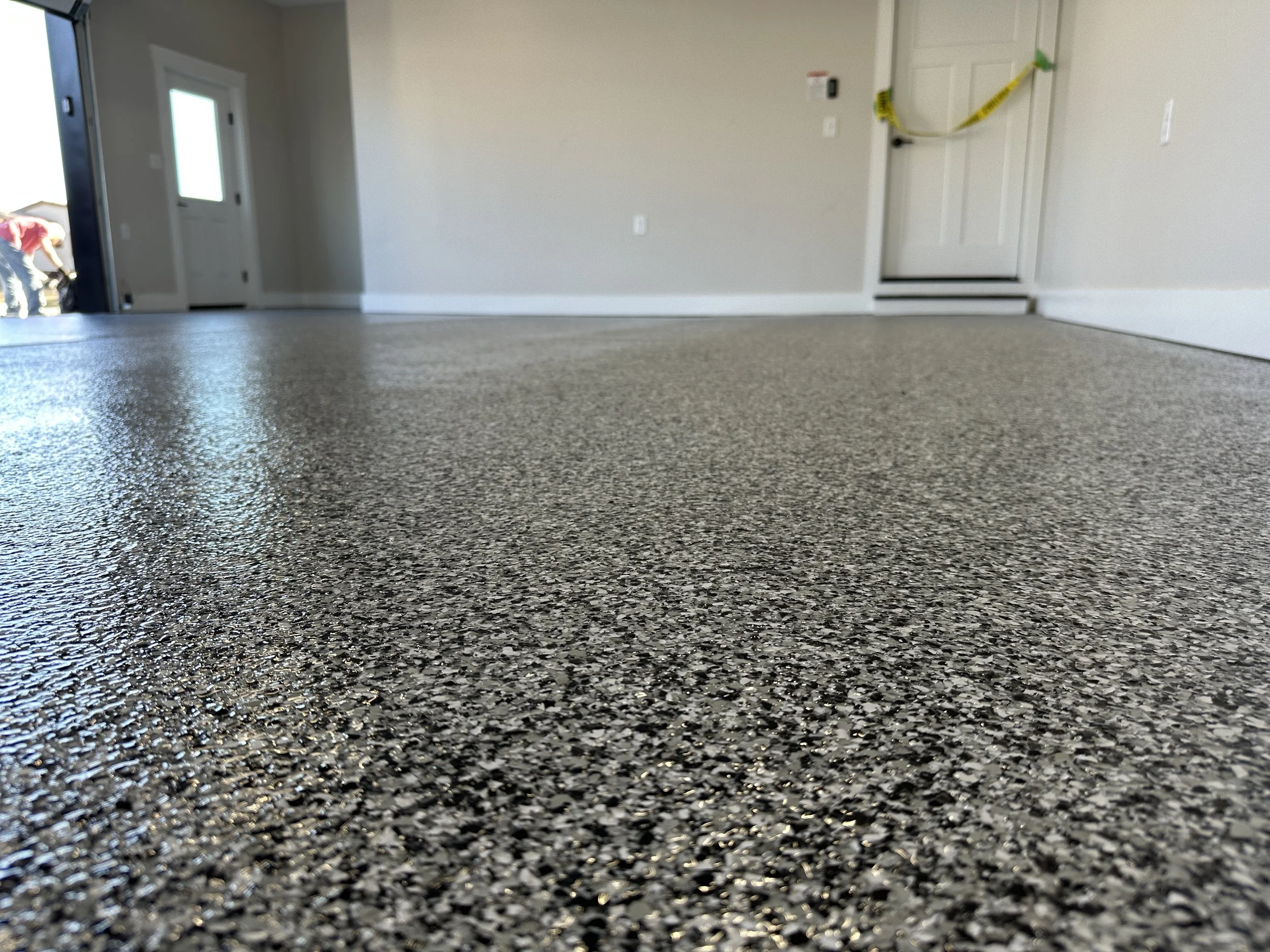 Close-up view of a textured gray epoxy or concrete floor in a room under construction, with a partially installed door and construction tape on the door in the background.