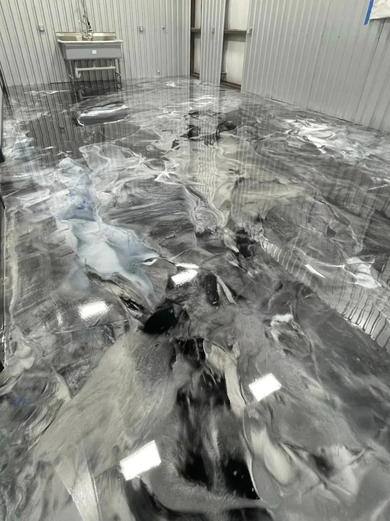 A room with a metal sink installed on the left and a metallic wall. The floor is covered with reflective, marbled black and white material, showing a wavy pattern.