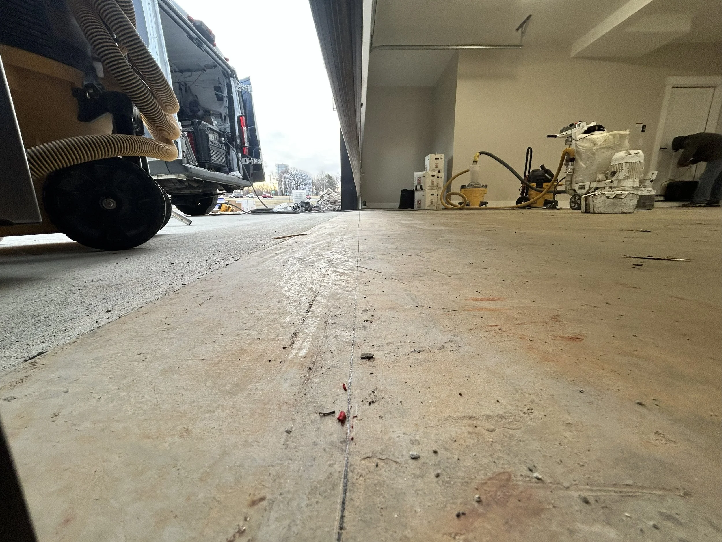 View of an indoor garage or workshop with construction or renovation work in progress. The floor has a strip of unfinished concrete, with various tools, equipment, and supplies scattered around. A person is working on something in the background.