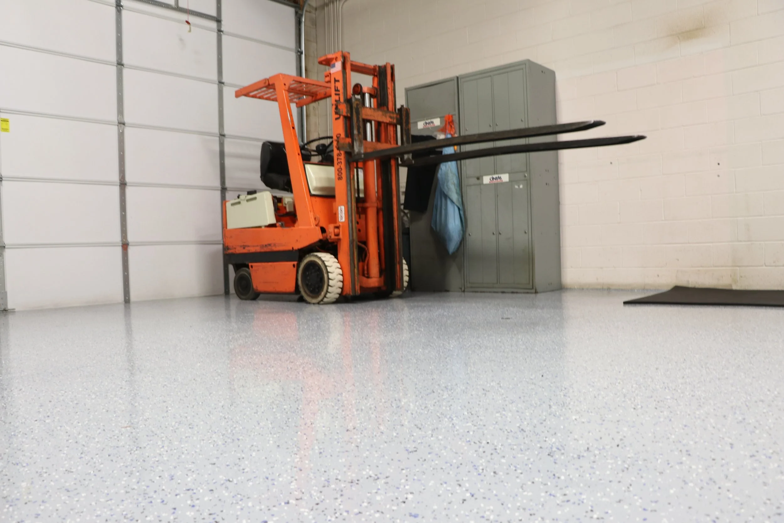 An orange forklift parked next to a gray storage cabinet in an indoor warehouse or garage with a light-colored speckled epoxy floor and a white brick wall.