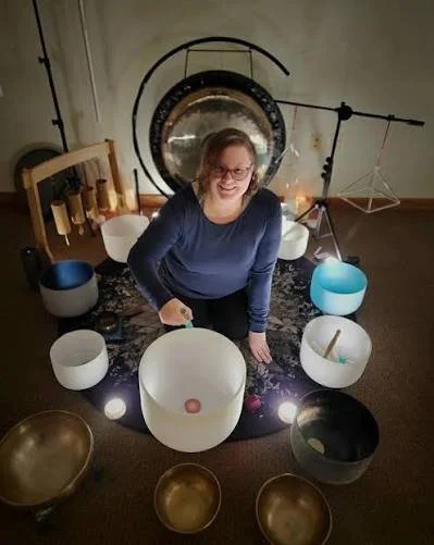 A woman with glasses sitting on the floor surrounded by large singing bowls and a gong in a room.
