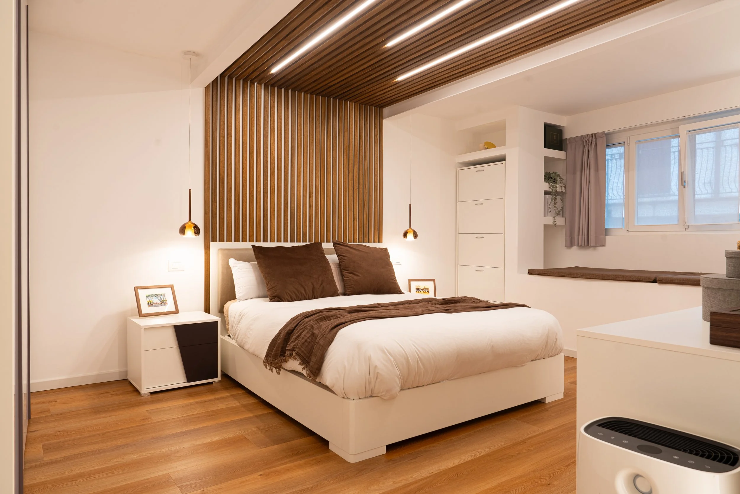 Camera da letto con letto matrimoniale, rivestimento in legno sul soffitto e sul muro alle spalle del letto, comodino bianco con una cornice, lampade a sospensione dorate, grande finestra con tenda beige, armadio bianco e una panca decorativa sotto l