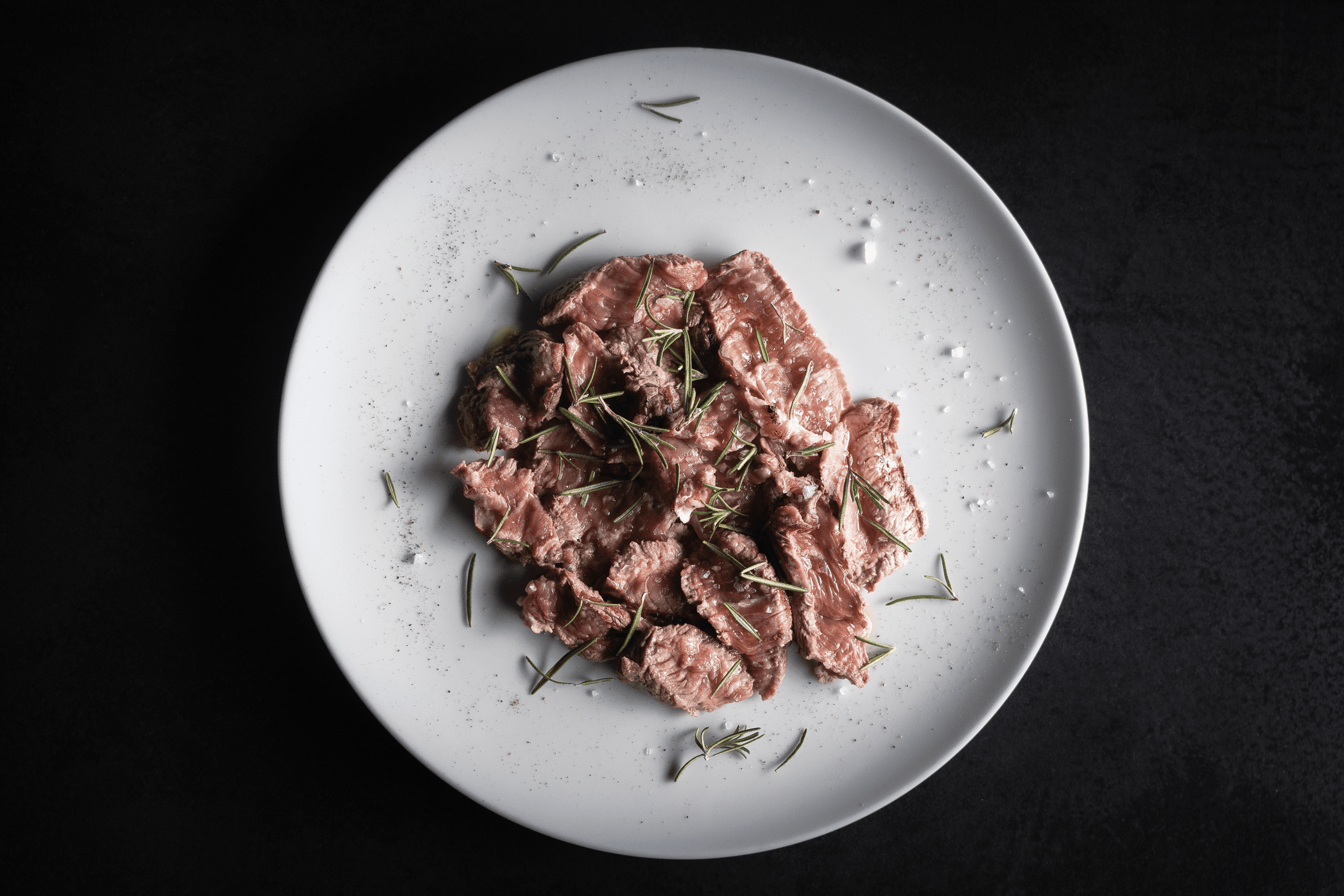 Fette di carne cruda condite con pepe, sale e rametti di rosmarino su un piatto bianco su sfondo nero.