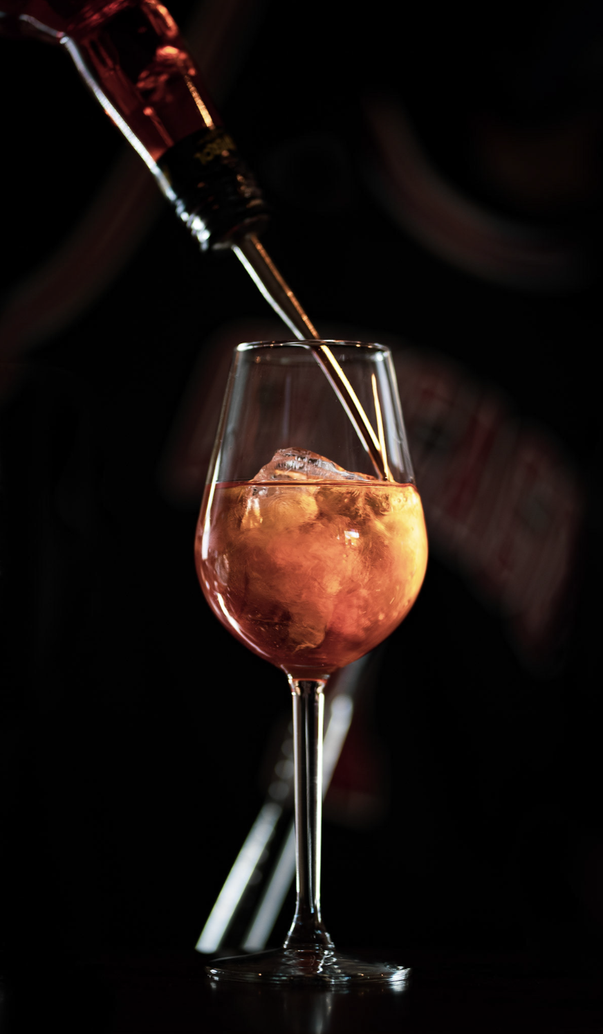 C Bermuda, un bicchiere da vino con una bevanda rosa, un cubetto di ghiaccio, mentre uno strumento di barrista versa una bevanda. Sfondo scuro