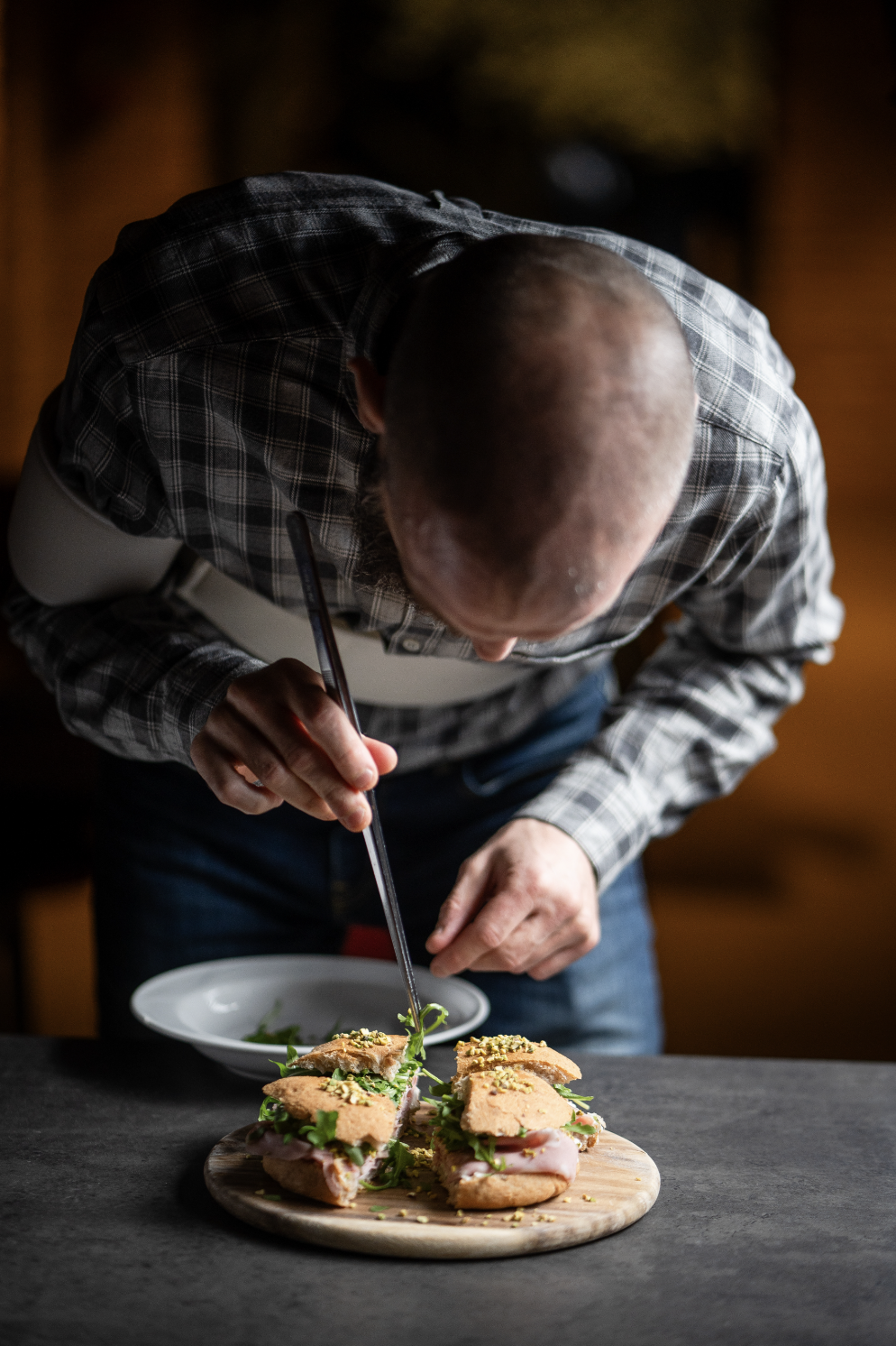 Uomo che prepara un panino gourmet, con ingredienti freschi e verdure, su un tagliere di legno.