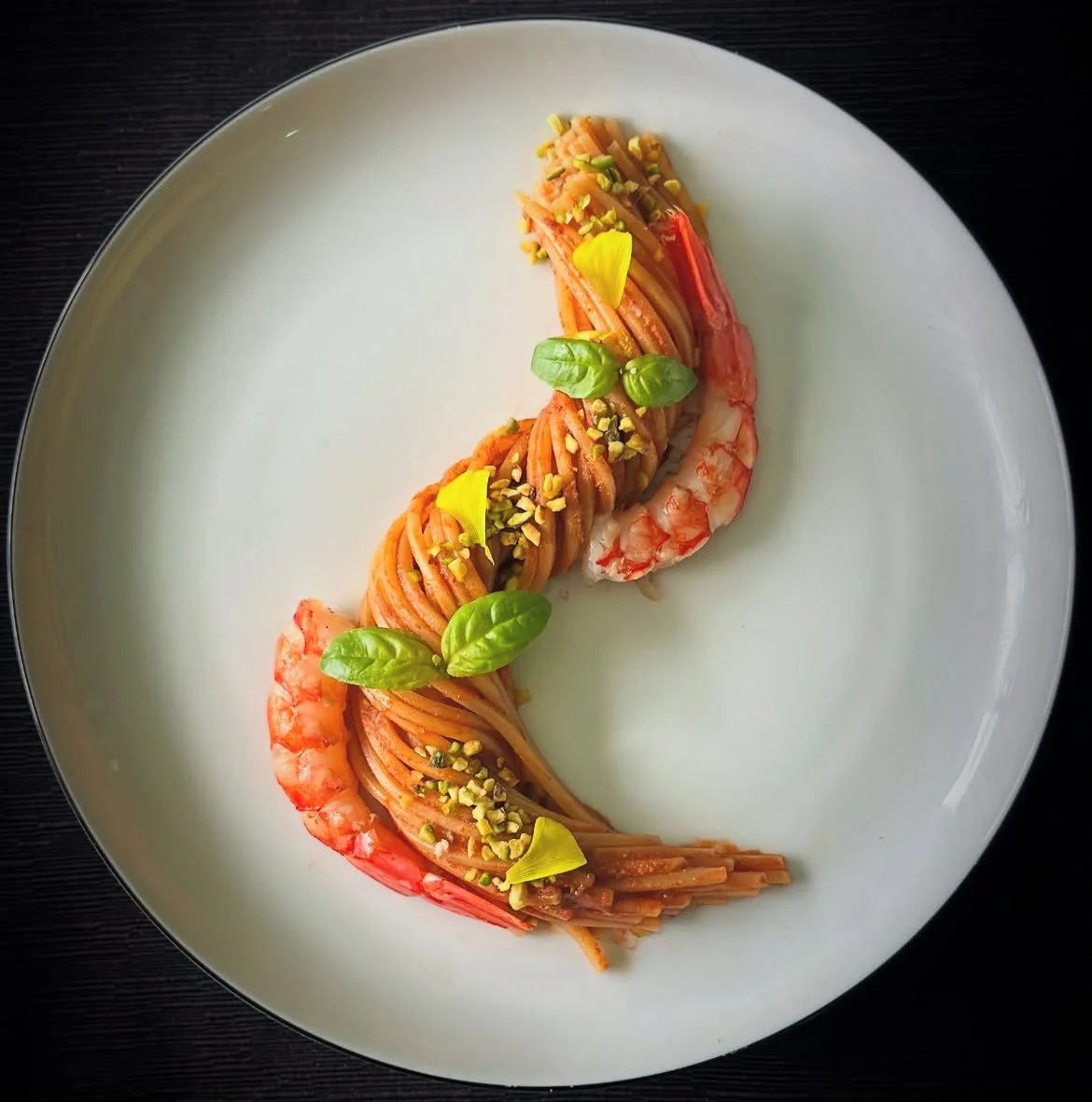 Piatto di spaghetti con gamberi, guarniti con foglie di basilico, pistacchi tritati e petali di fiori gialli, serviti su un piatto bianco.