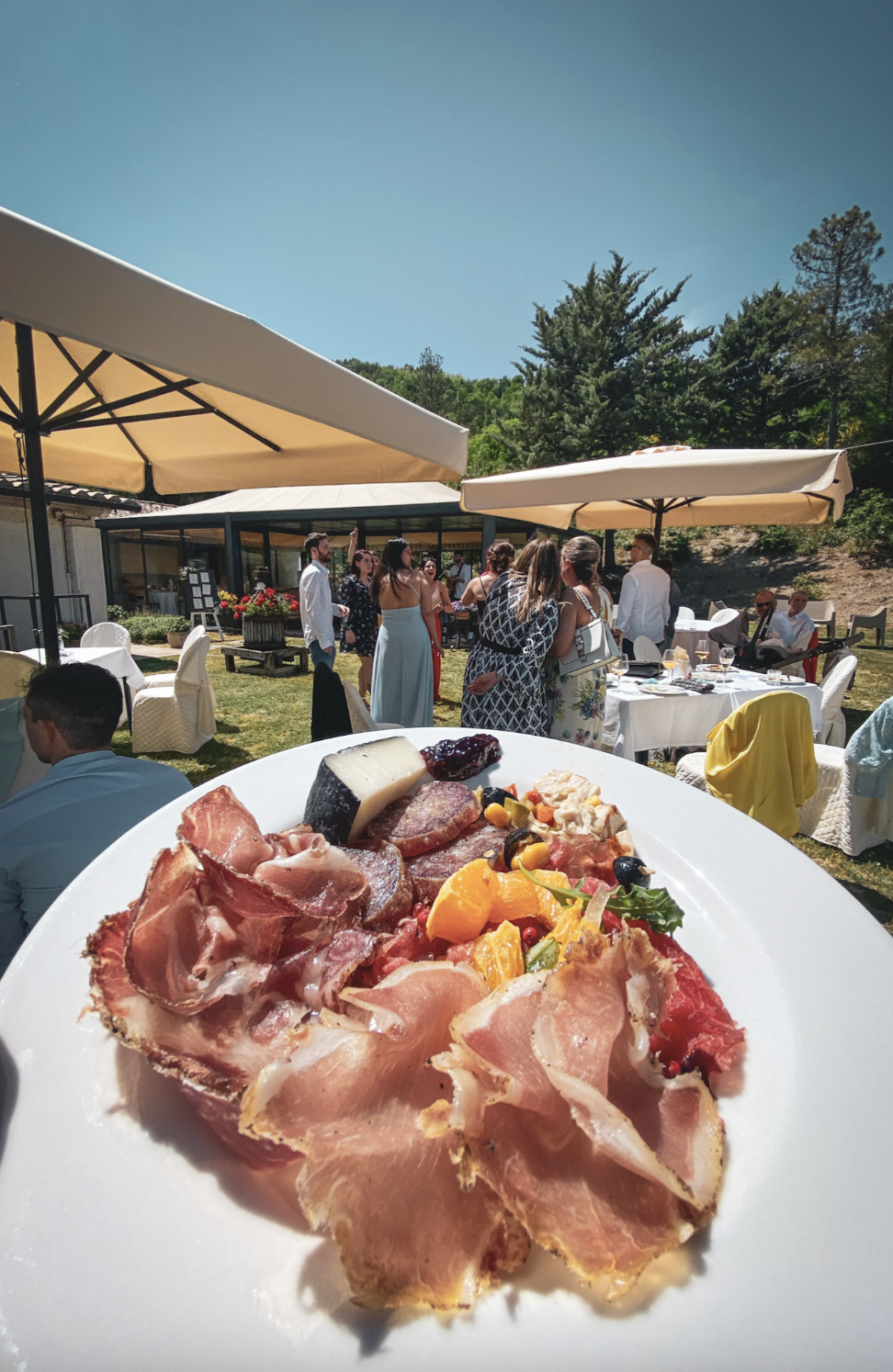 Un piatto di antipasti con prosciutto, formaggi, frutta e olive, con un gruppo di persone che si socializza all'aperto durante una festa o un matrimonio