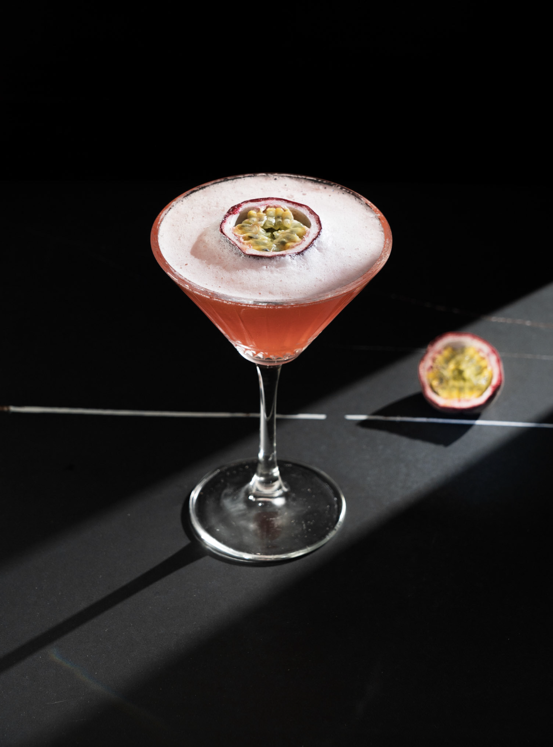 Un cocktail rosa in un bicchiere da martini con una guarnizione di passione, accanto una metà di frutto della passione su uno sfondo nero.