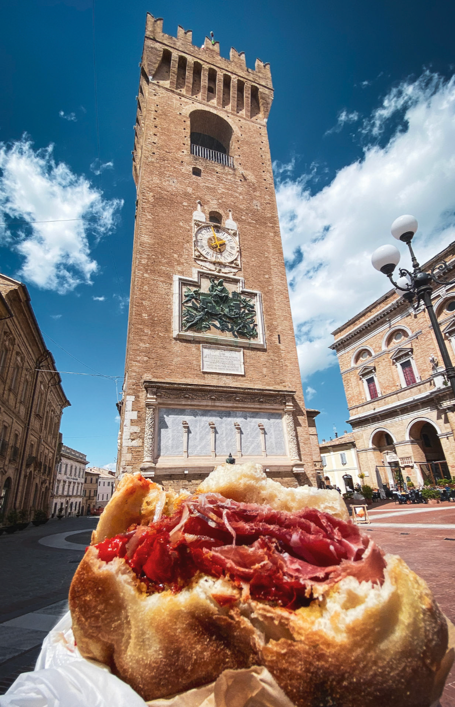 Pani con prosciutto crudo e pomodori in primo piano, Torre dell'Orologio in Piazza dell'Erbe a Verona sullo sfondo con cielo sereno.