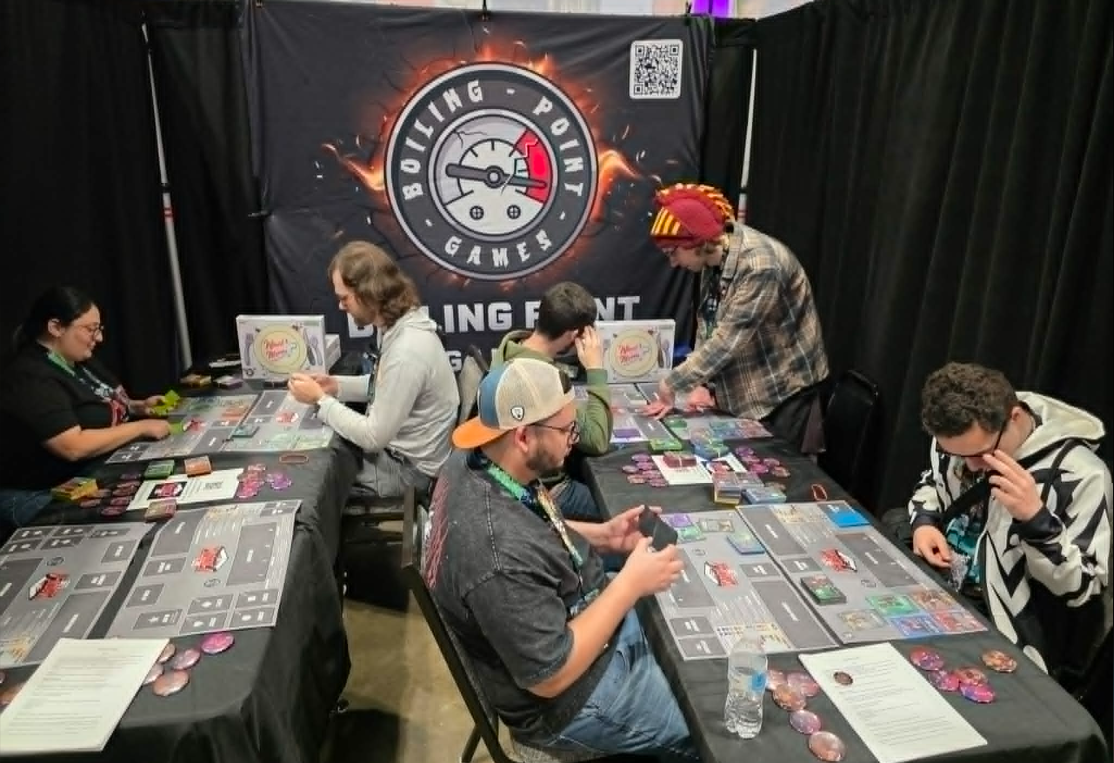 People playing a board game called 'Ready Set Fight' at a gaming event, with a large wall banner featuring Boiling Point Game's logo behind them.