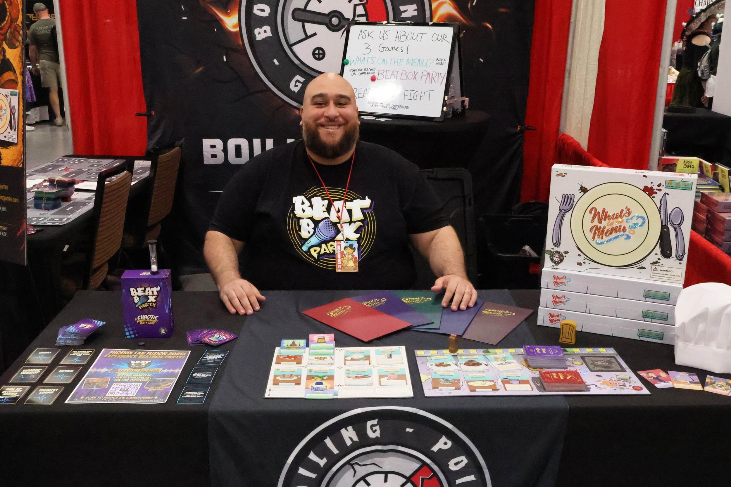 A man with a beard and a big smile sitting at a Boiling Point Games table at a gaming event, with game boxes, cards, and promotional materials for games called Beat Box Party and What's On The Menu displayed on the table.